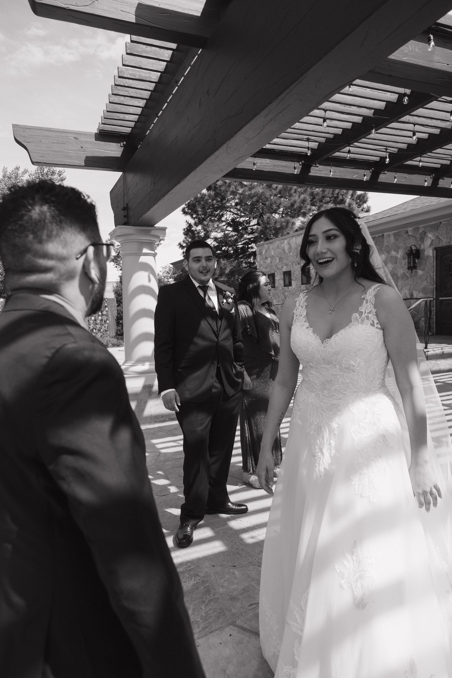 Black and white photo of smiling bride walking toward groom under pergola with wedding guests nearby.