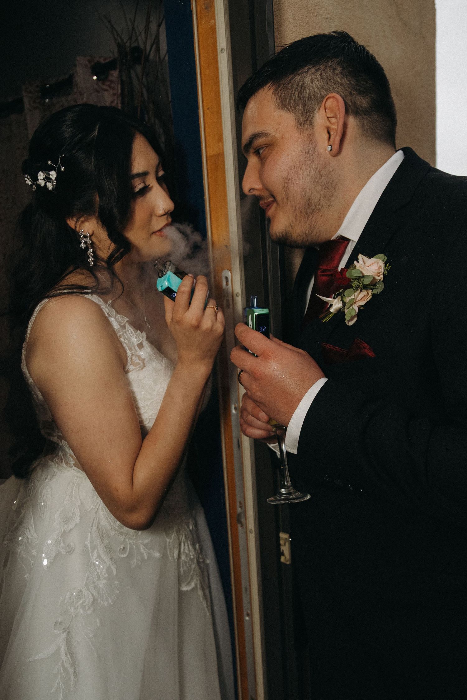 Bride and groom share an intimate moment near a doorway, with the bride holding a colorful vape device.