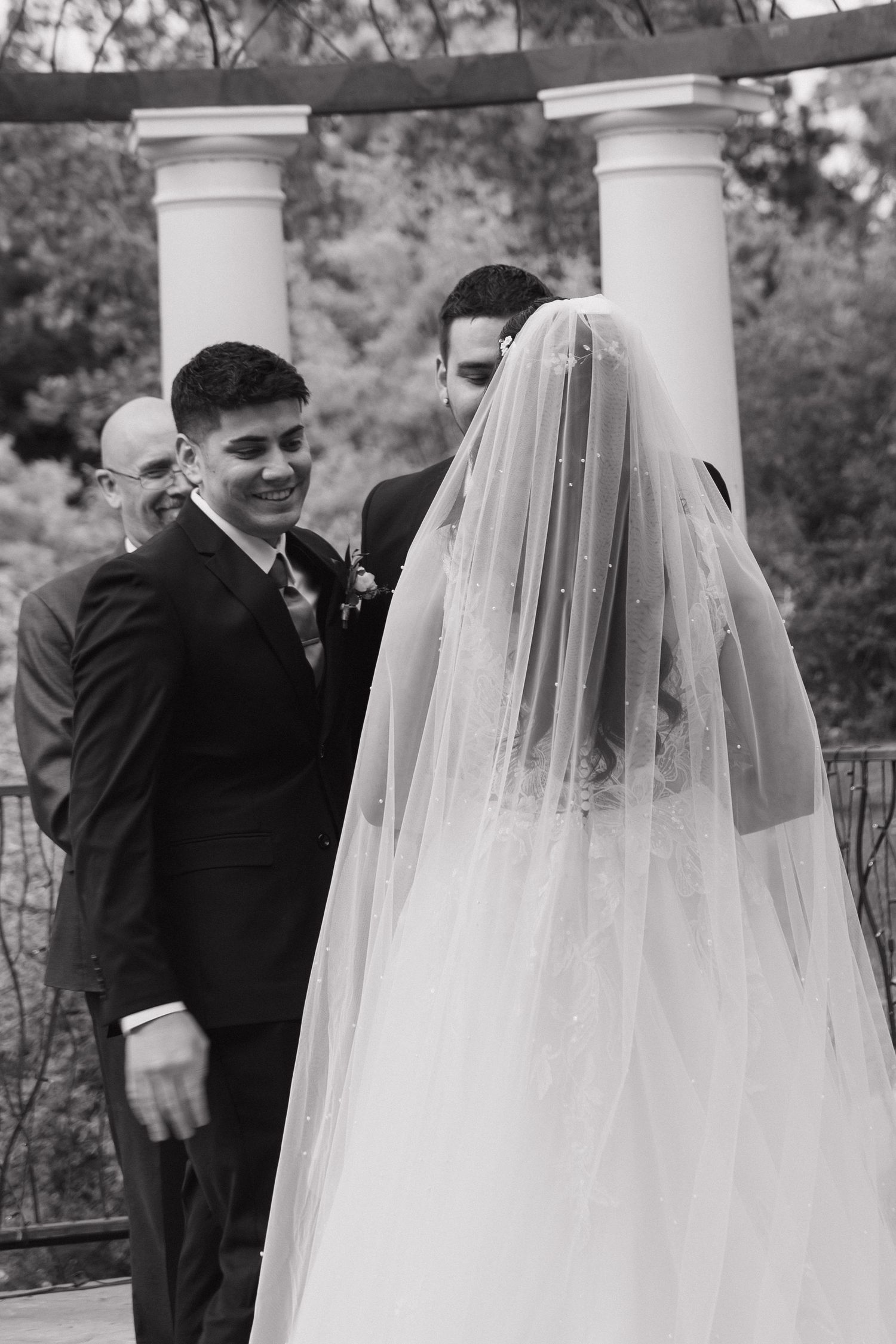 Black and white photo of smiling groom watching veiled bride approach at outdoor wedding ceremony with columns.