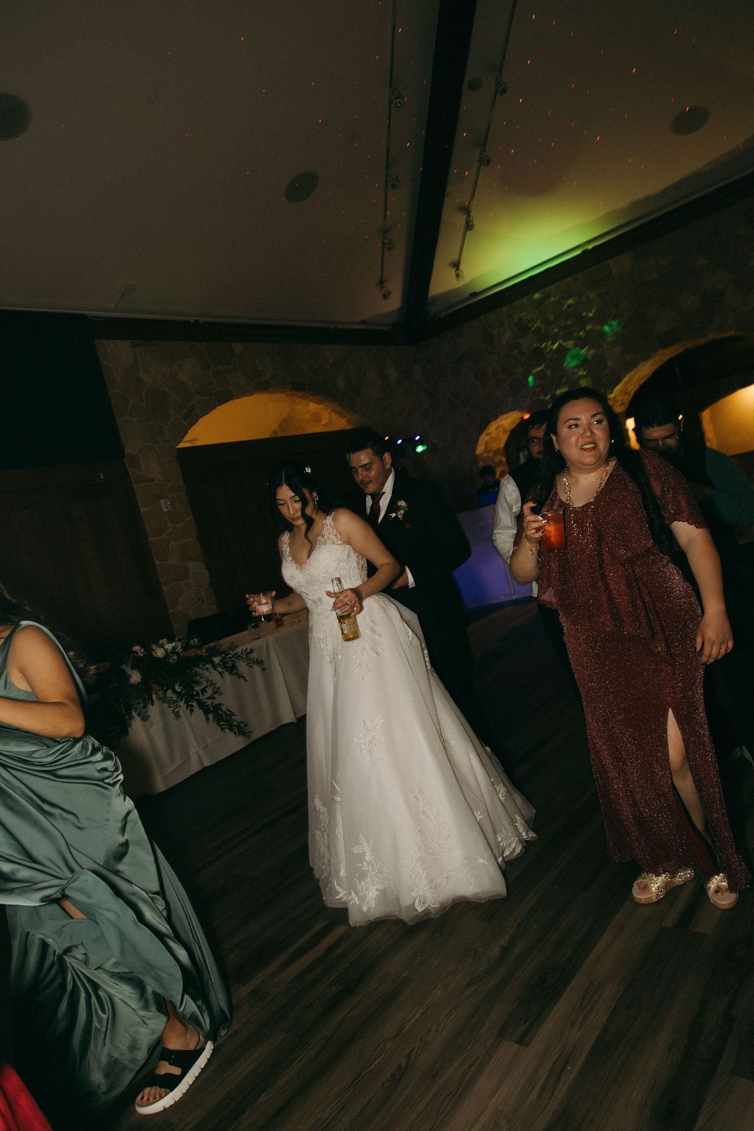 Bride in white ball gown dances with groom and guests under colorful lights at wedding reception.