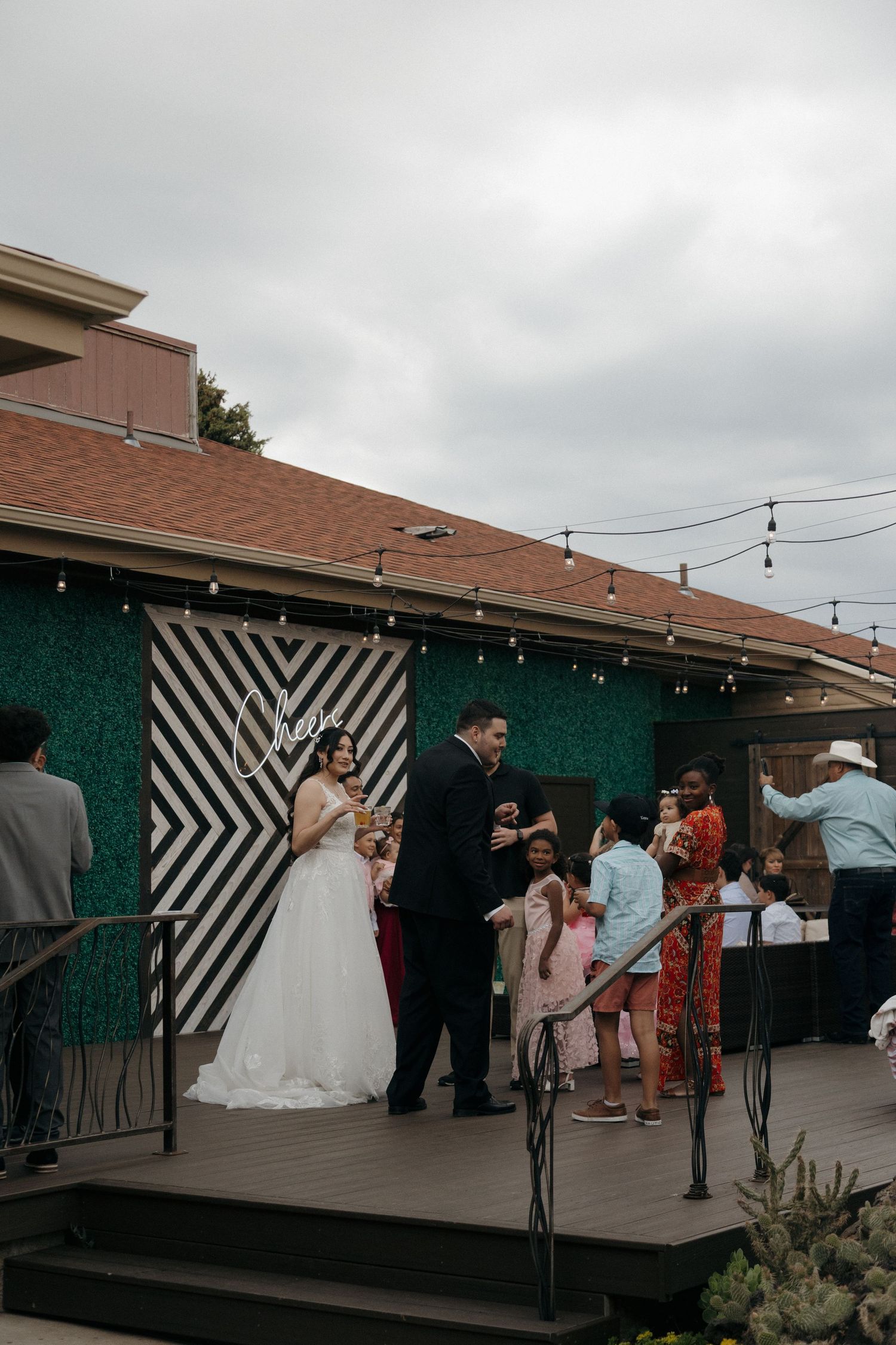 Bride and groom celebrate on outdoor deck at wedding reception with guests and string lights.