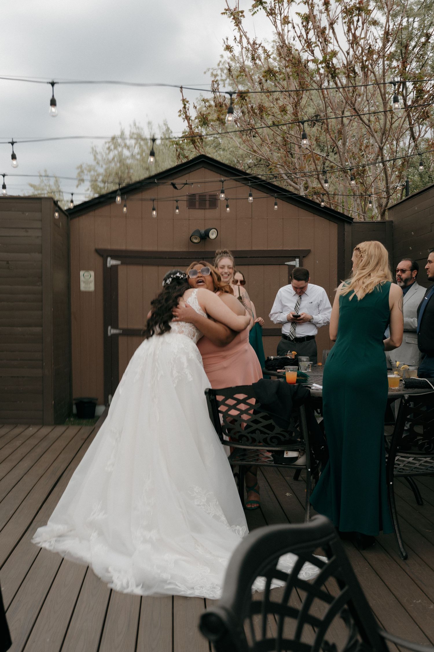 Bride in white gown hugging guest in pink dress at outdoor wedding reception on wooden deck.