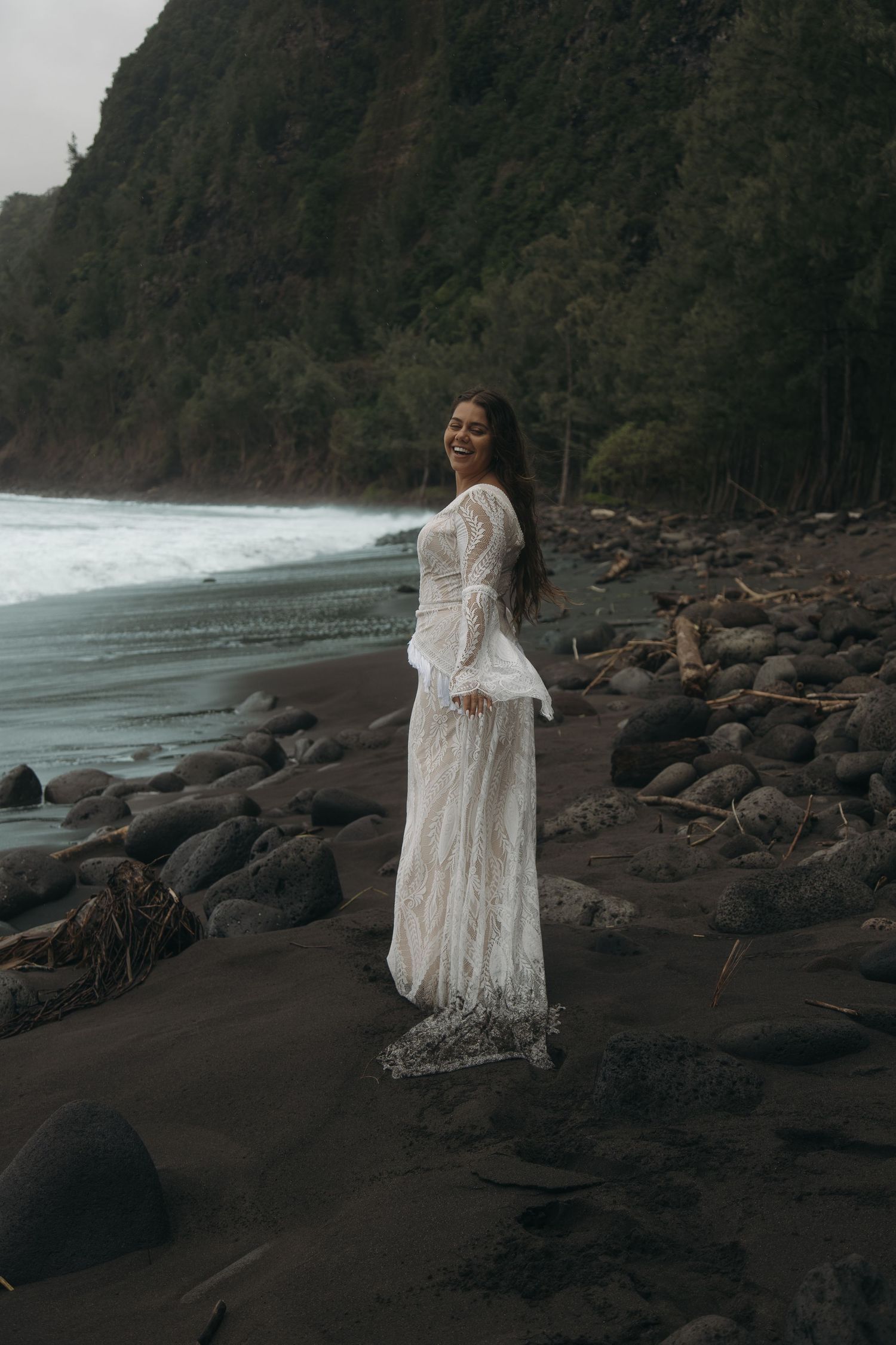 Bride in white boho lace bell-sleeve gown smiles on rocky black sand beach with green cliffs.