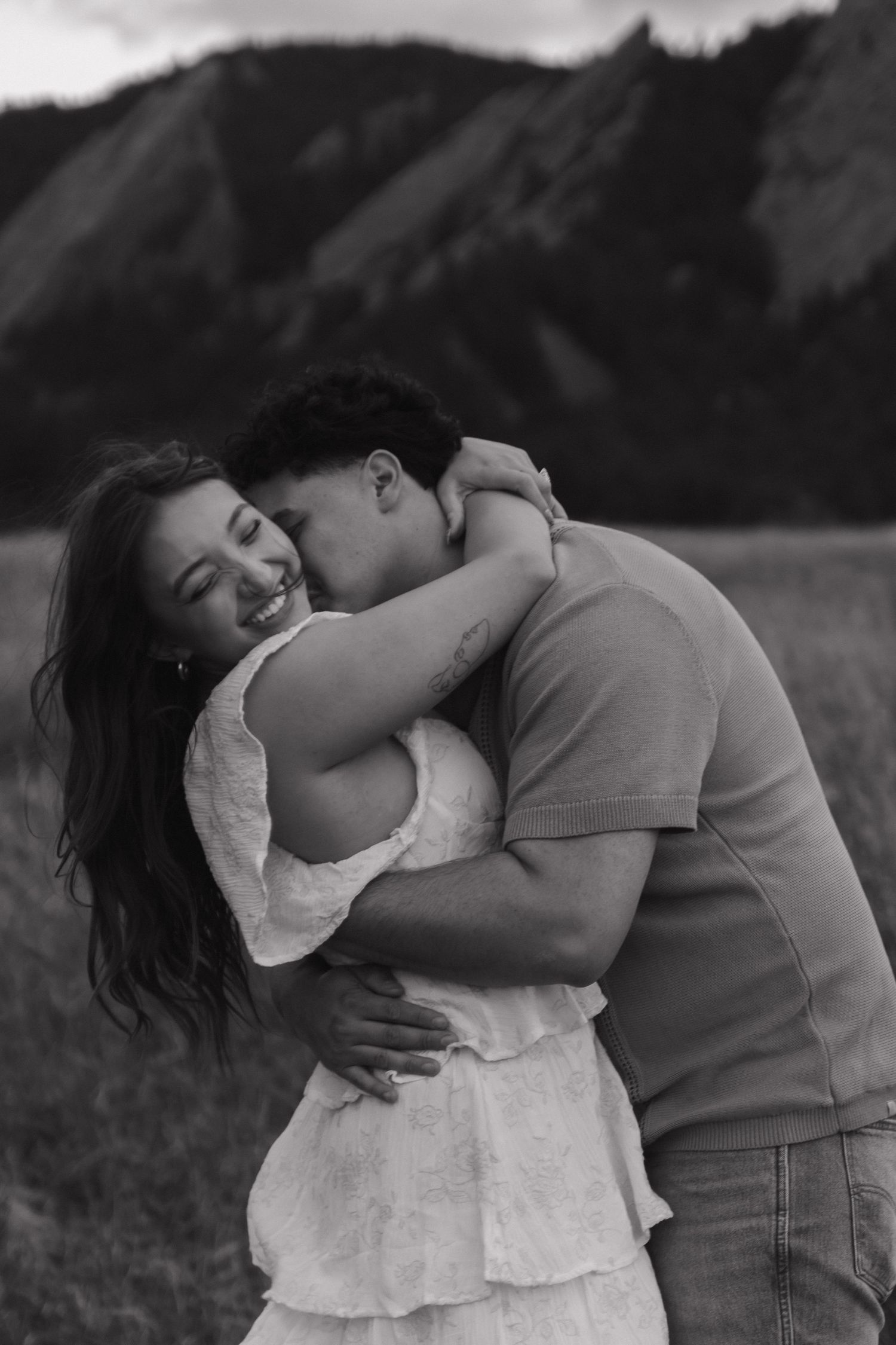 A black and white photo of a man embracing and kissing a laughing woman near a mountain backdrop.
