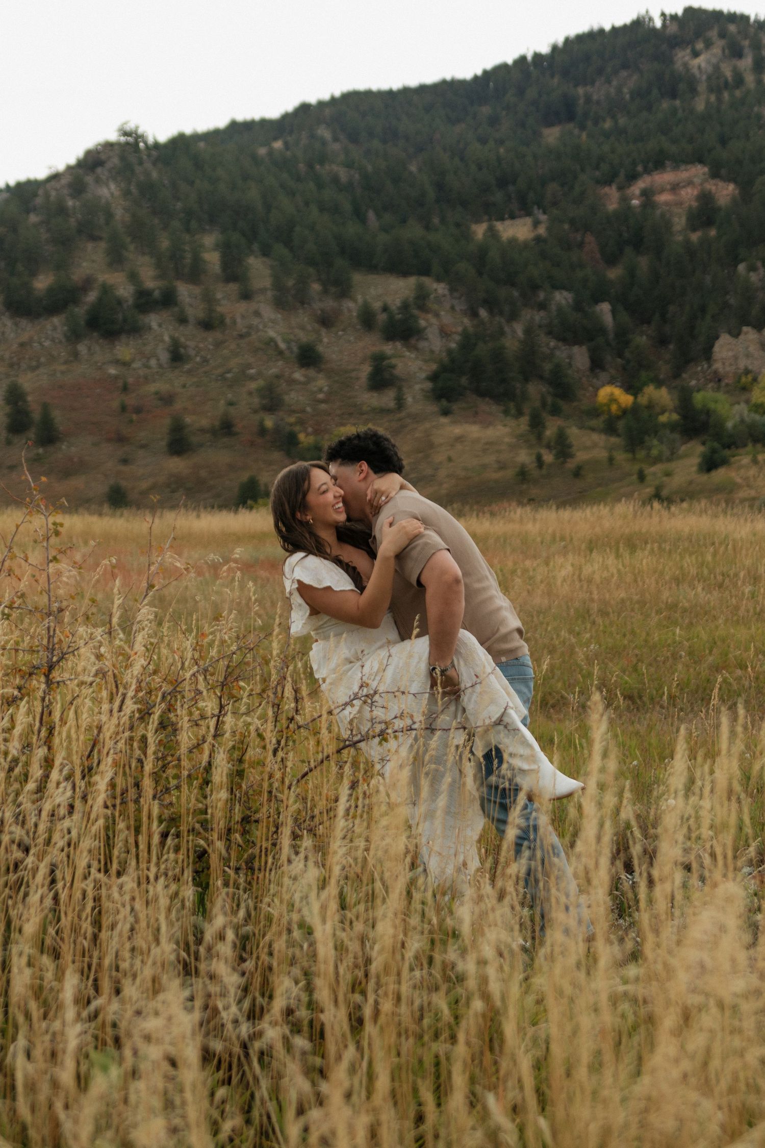 Romantic couple kissing in tall golden grass with a forested mountain backdrop.