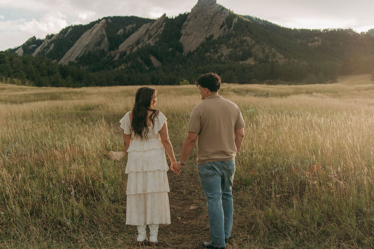 Couple holding hands walking toward rocky Flatirons mountains through golden grass field.