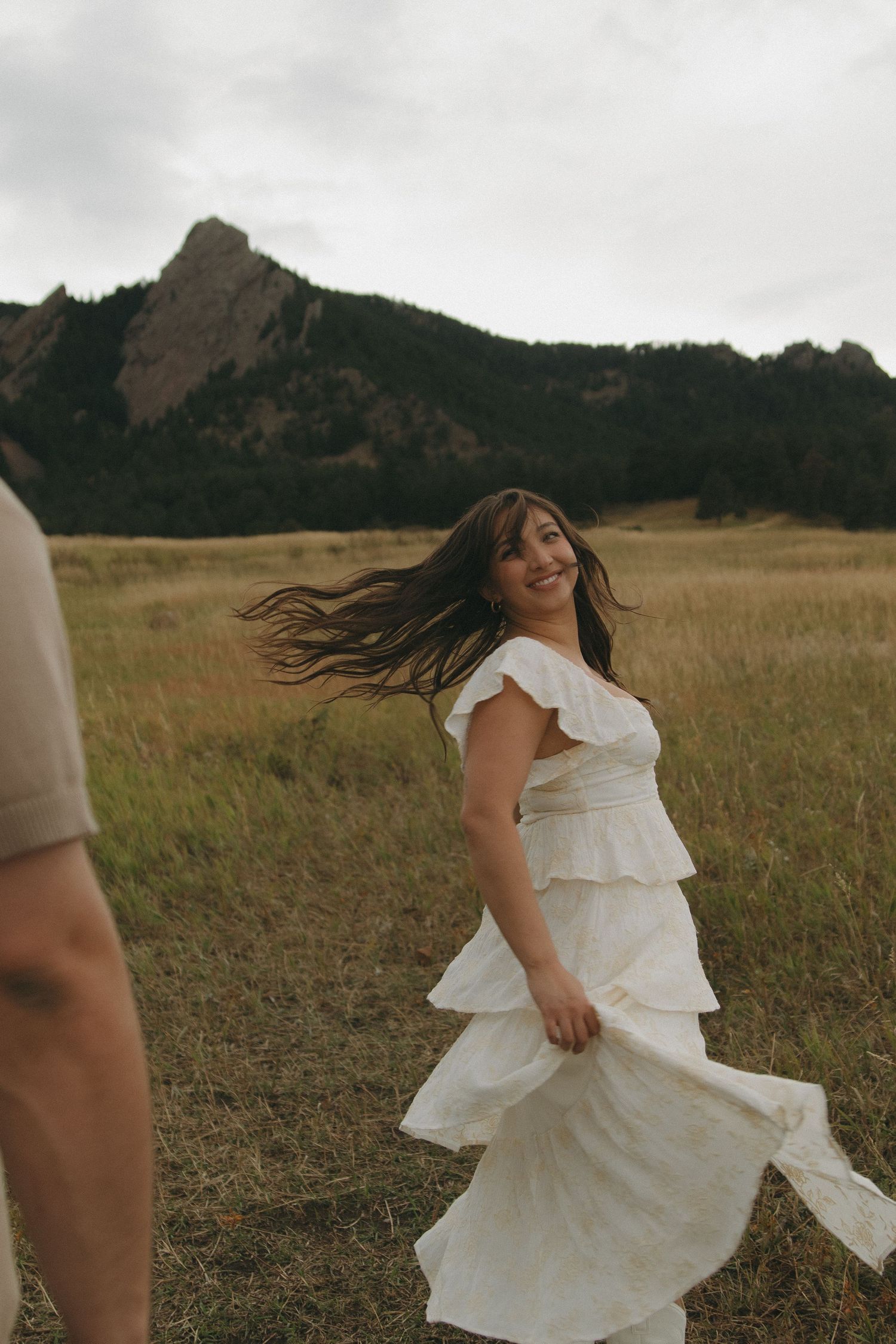 Woman in white ruffled dress twirls and smiles in a meadow with the Flatirons in the background.