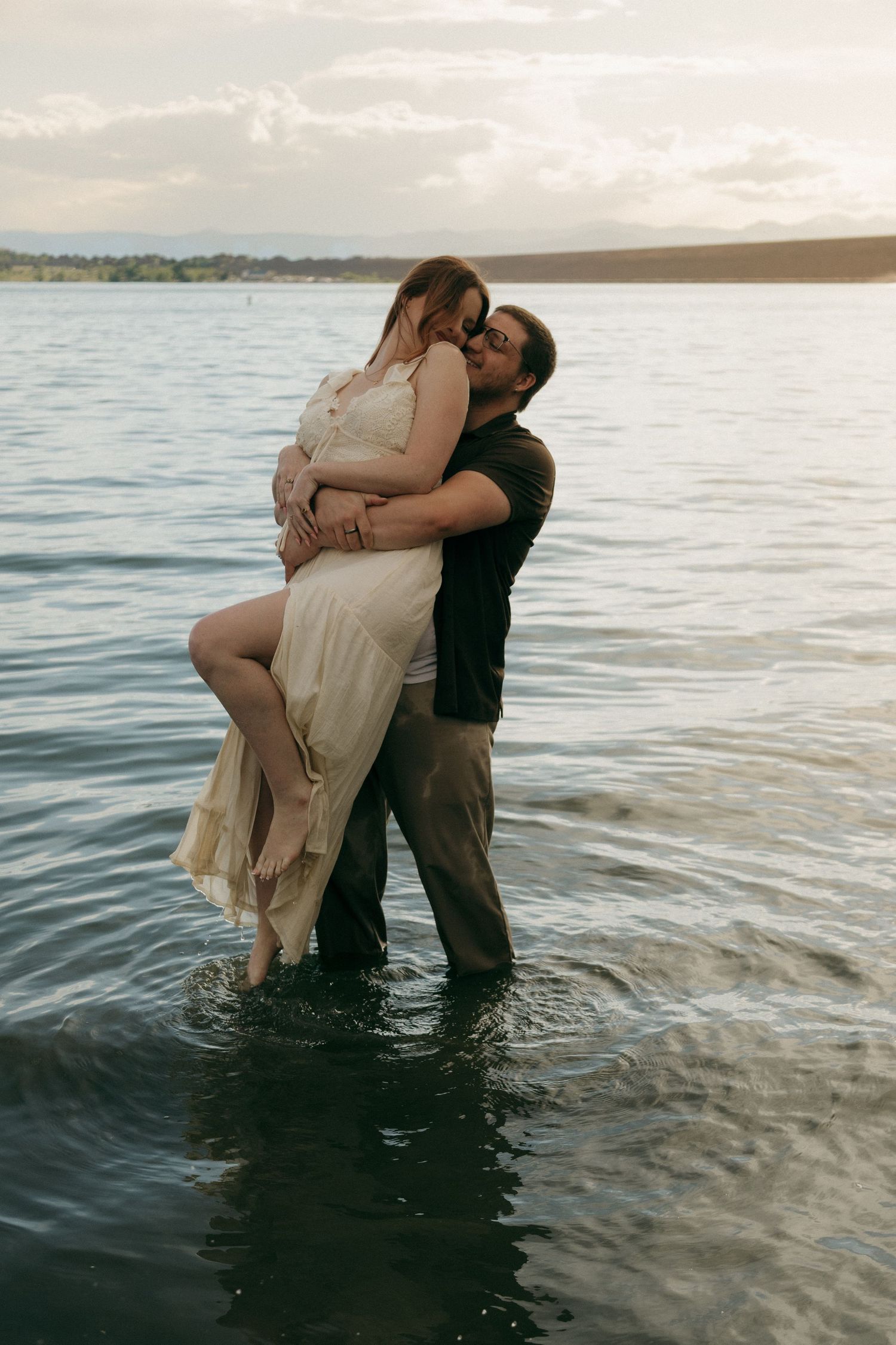 Romantic couple embracing in lake water at sunset, man lifting woman in cream lace dress.