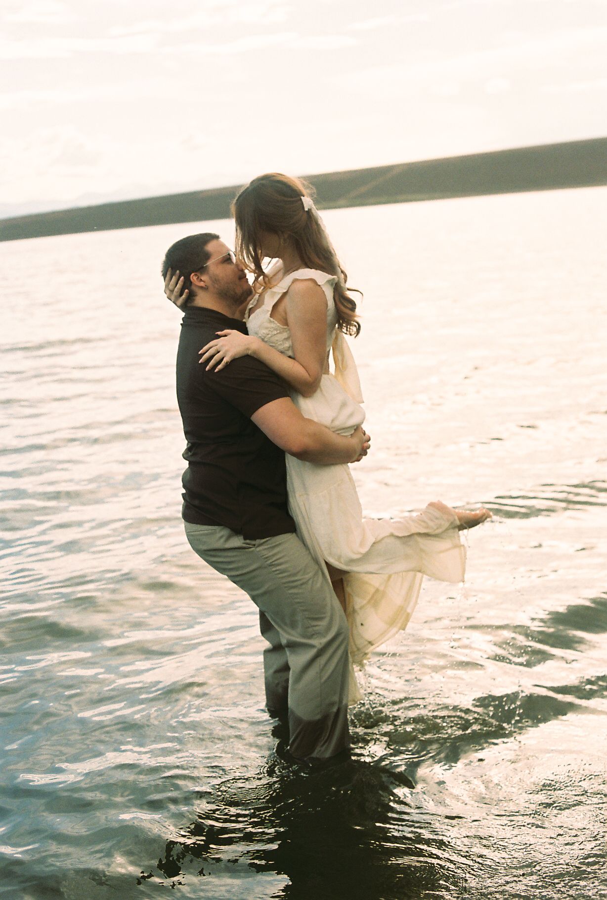 Romantic couple embracing in shallow lake water at golden hour, man lifting woman in white dress.