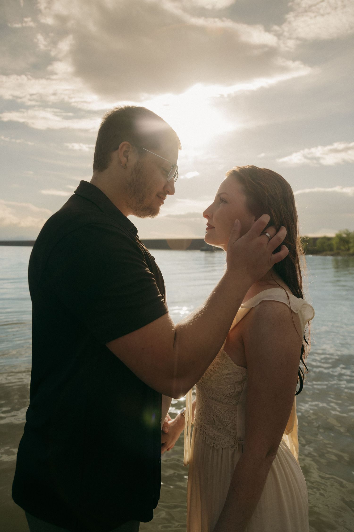 Couple gazing into each other's eyes near a lake with warm sun flare and cloudy sky backdrop.
