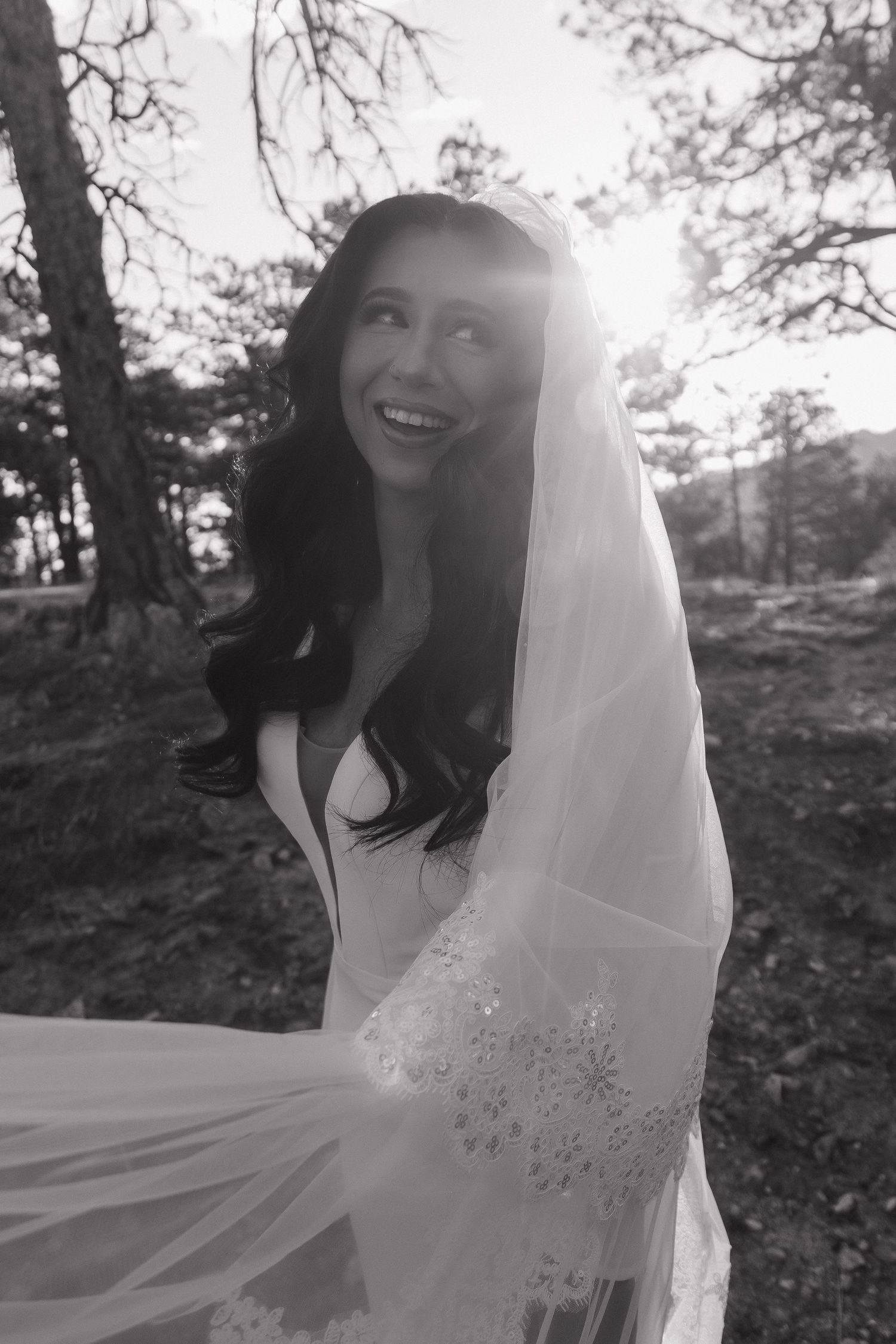 Black and white photo of smiling bride with flowing veil backlit by sunlight in forest setting.