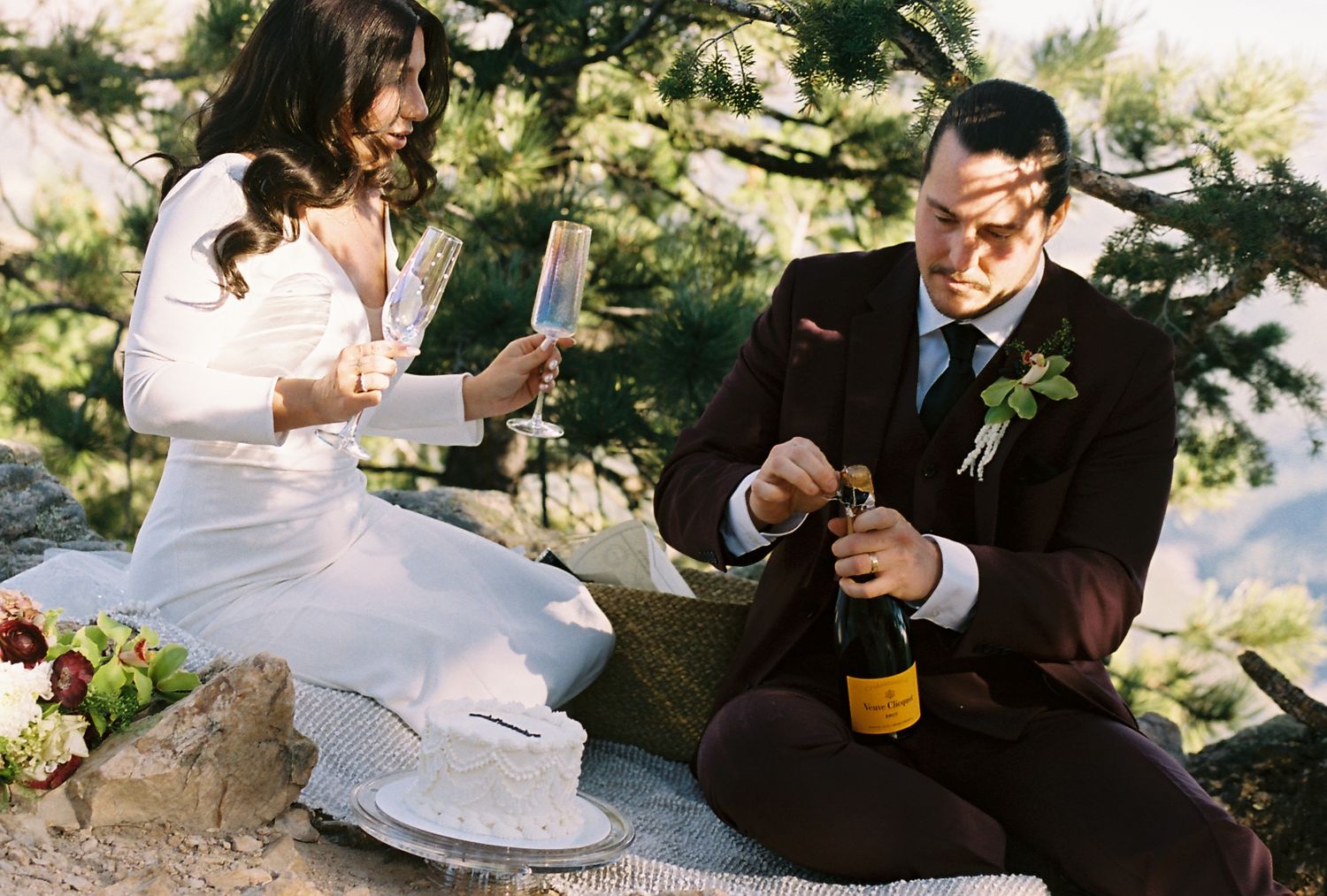 Bride reads vows while groom opens champagne bottle as they sit on a rocky ledge at outdoor elopement.