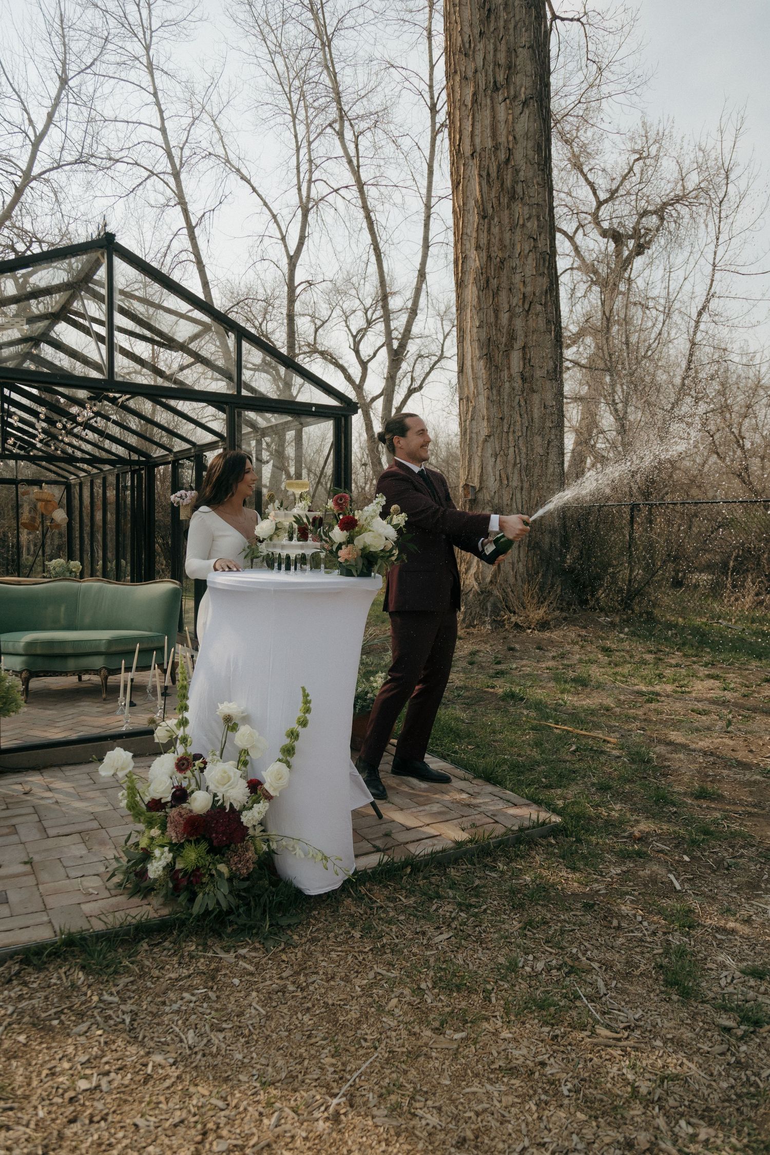 Groom sprays champagne bottle outdoors near a white cocktail table with floral arrangements at wedding celebration.