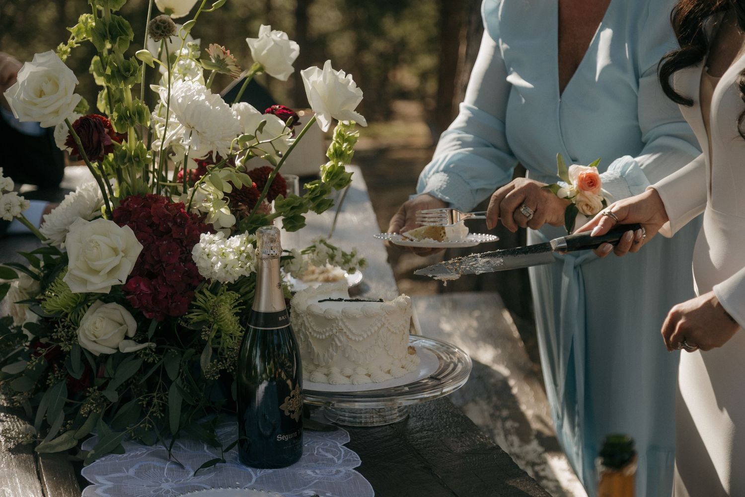 Wedding cake and champagne bottle on table with white and burgundy floral arrangement outdoors.