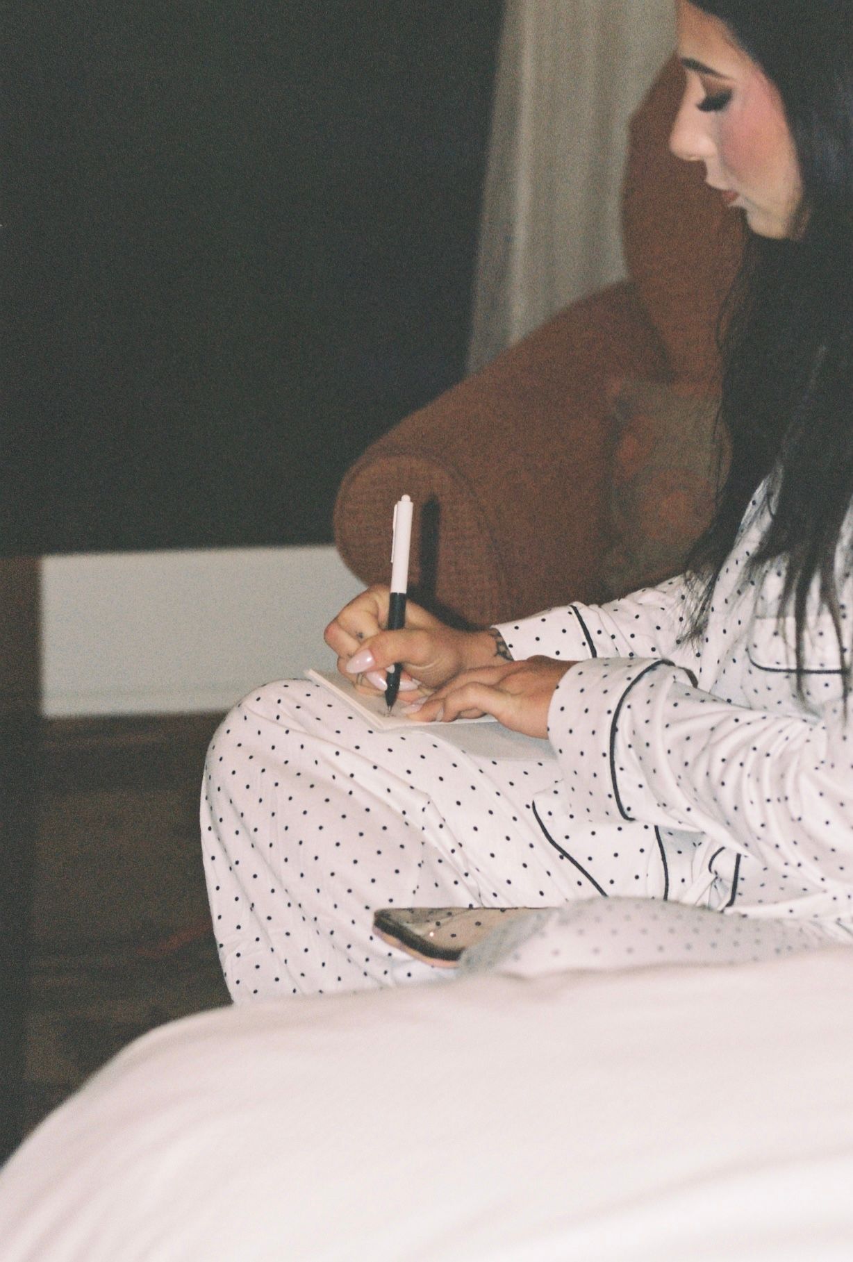 Bride in polka dot pajamas writing vows on bed in softly lit room before wedding day preparations.