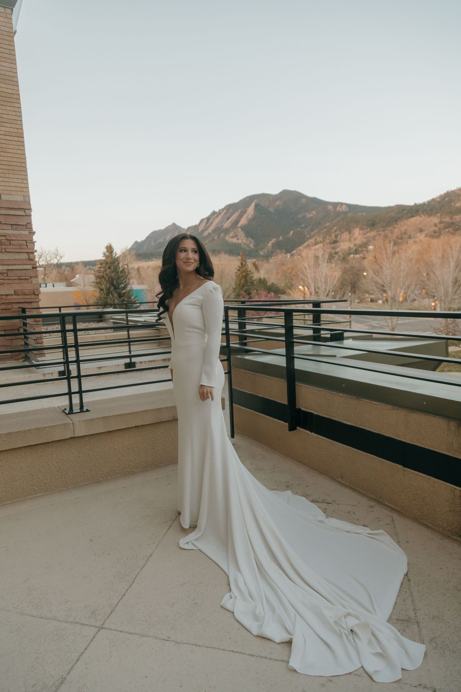Bride in a long-sleeve white wedding gown with train poses on balcony with mountain views.