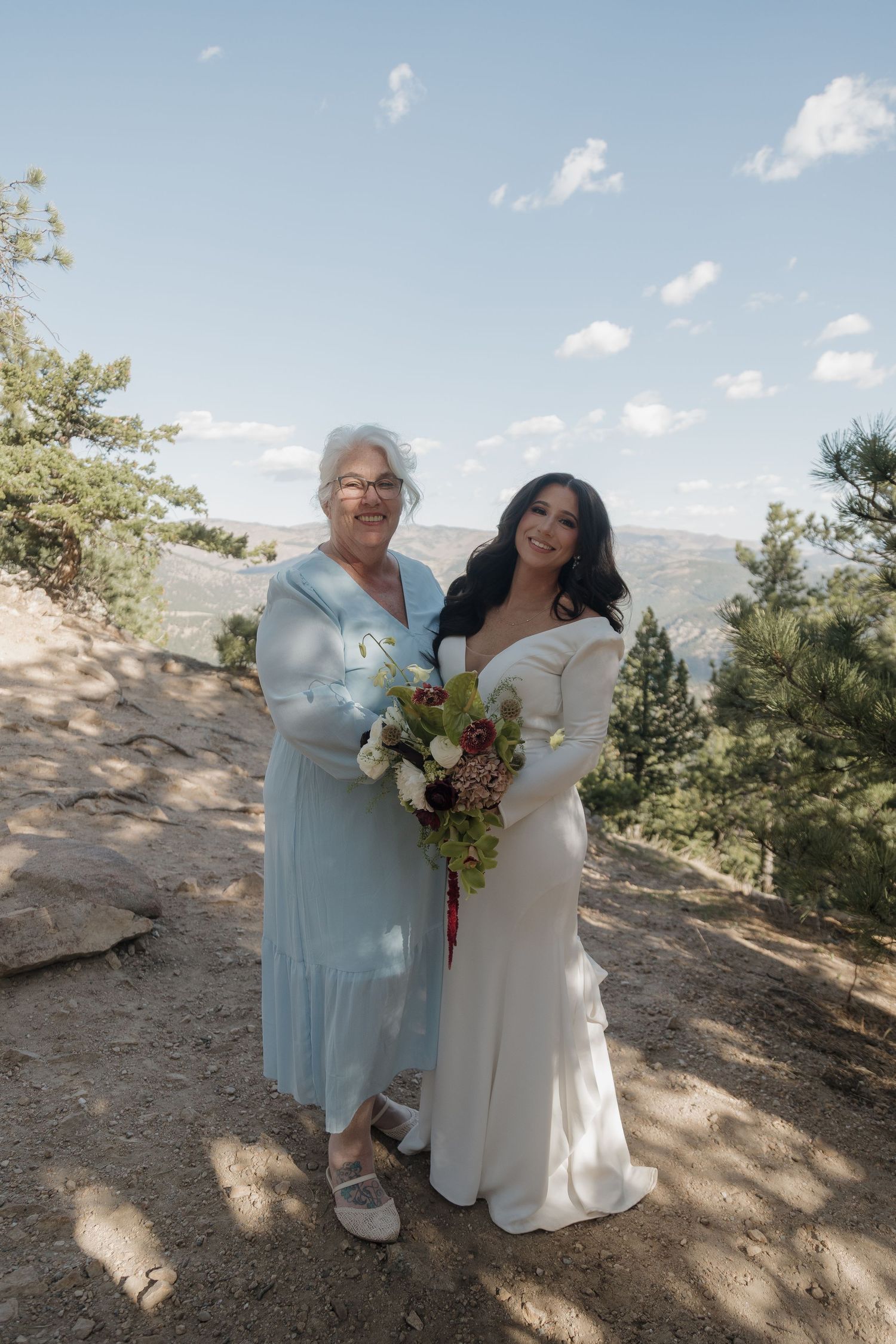 Bride in white off-shoulder gown poses with older woman in blue dress holding bouquet on mountain overlook.