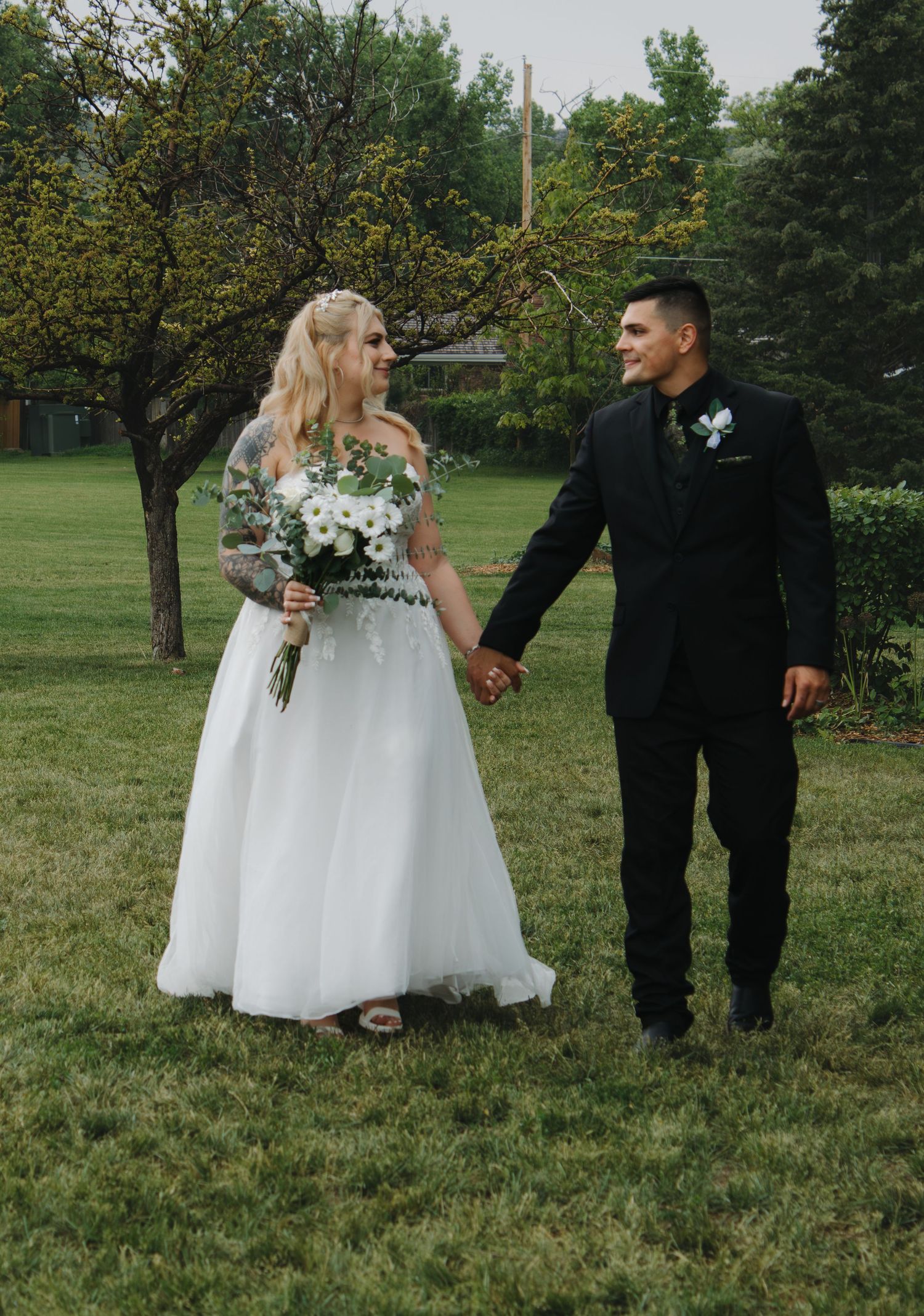 Couple in wedding attire walking through a lush green garden setting.