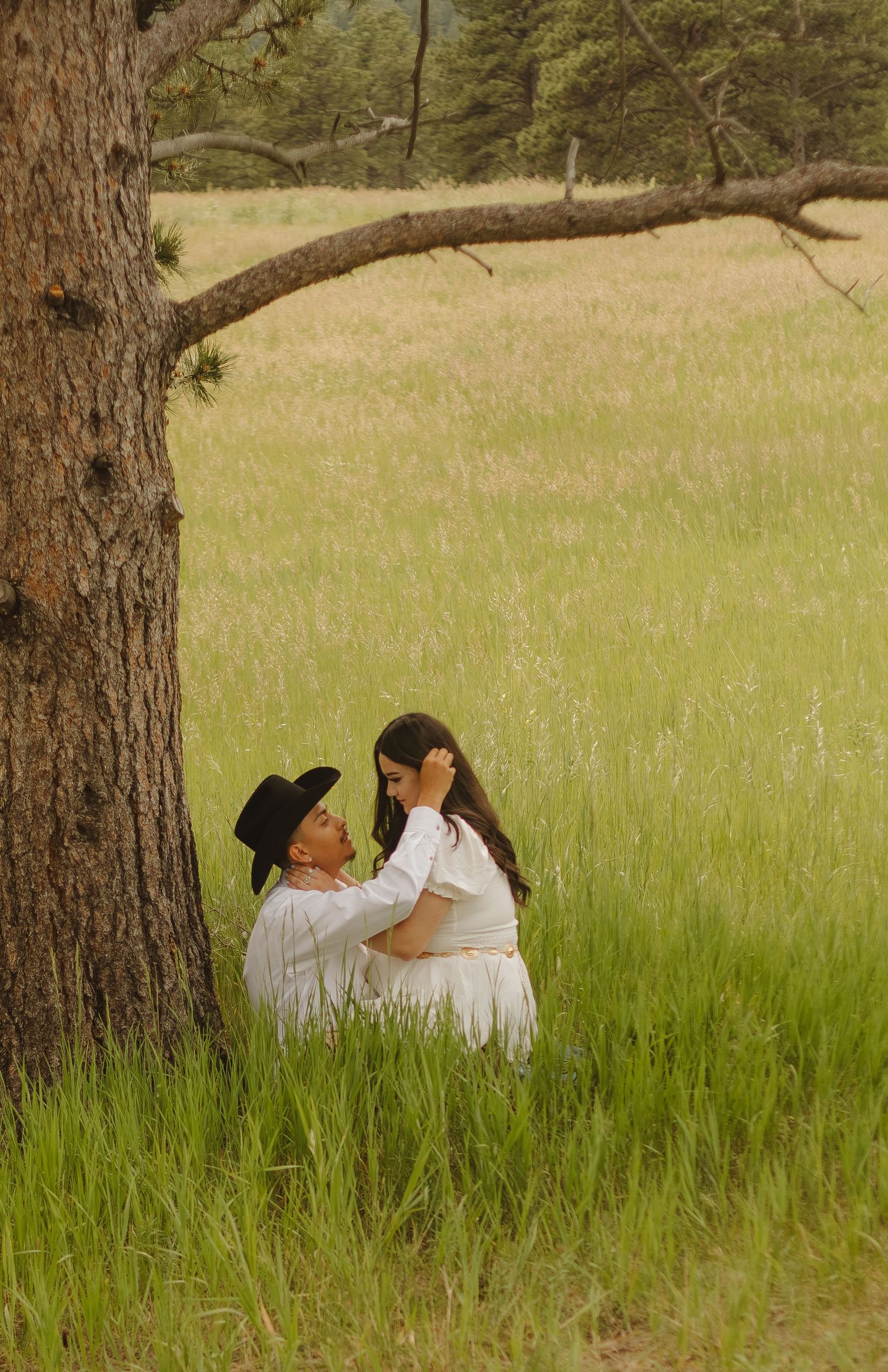 A romantic couple in light colored clothing sharing a sweet moment by a tree in a grassy field.