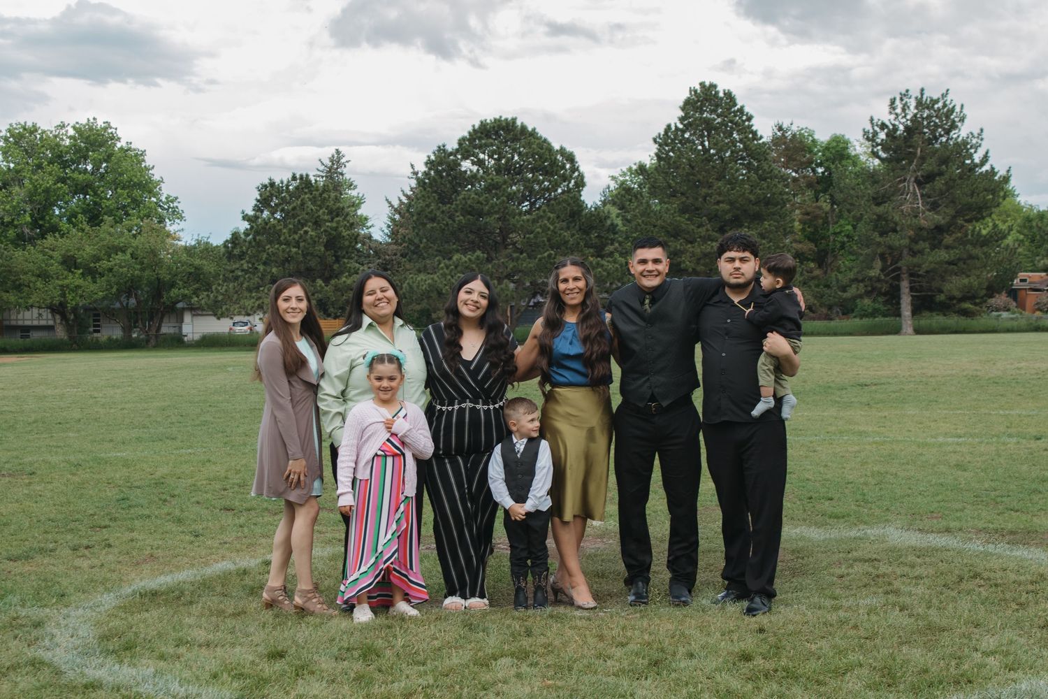 Group photo of wedding guests standing outdoors on grassy field with trees in background.