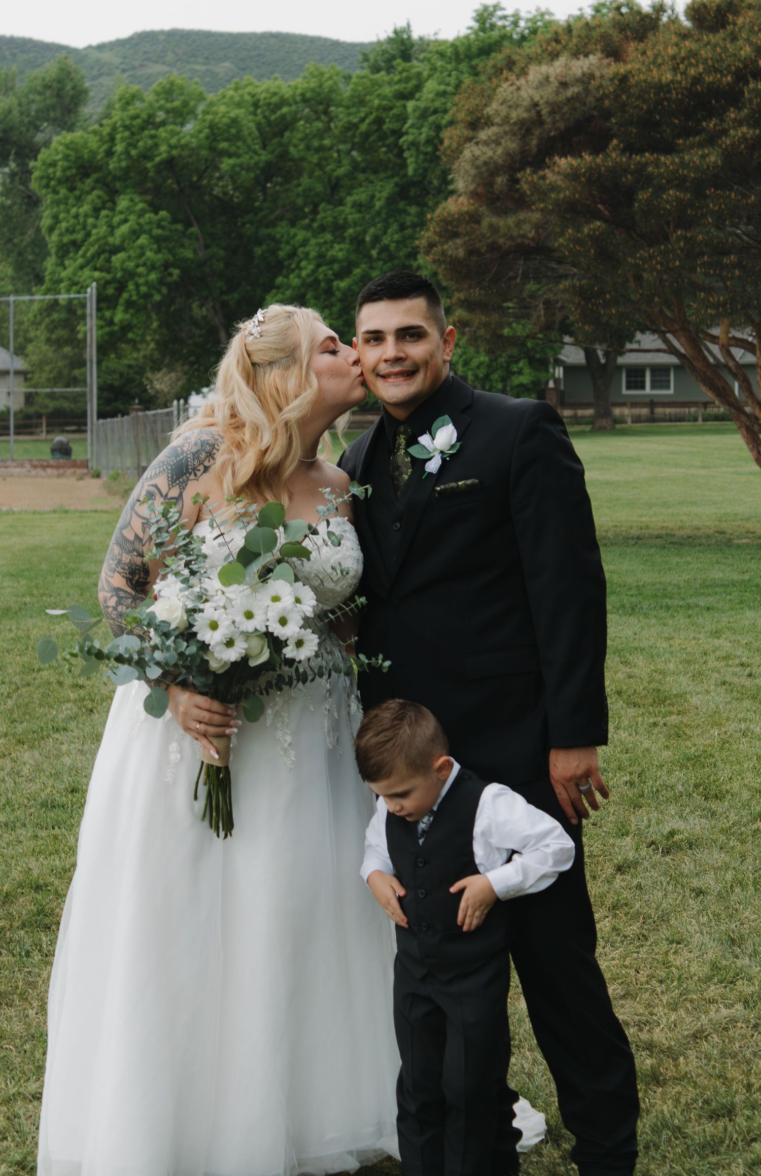 Candid wedding moment with bride and groom interacting with young ring bearer outdoors.