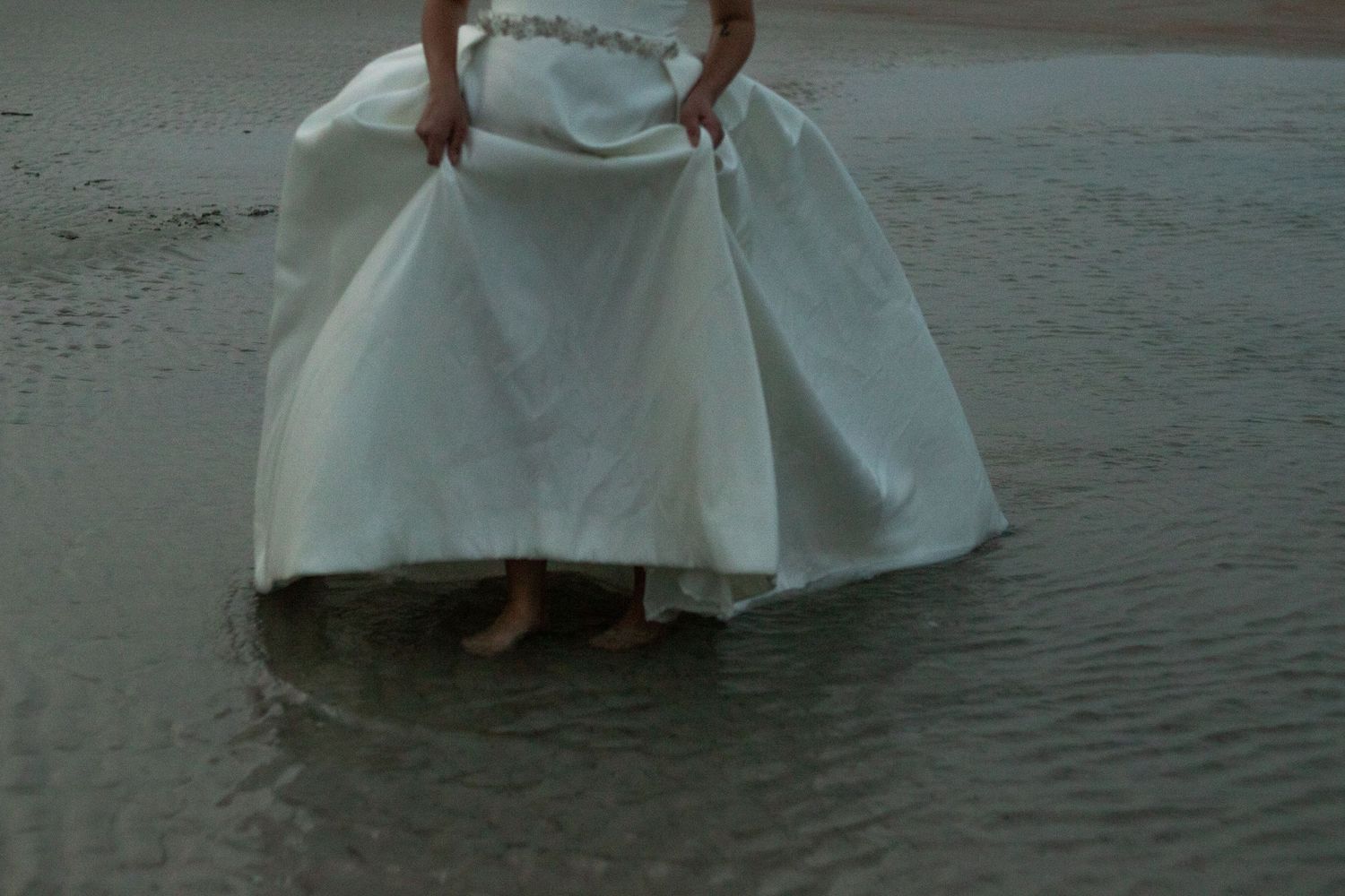 The hem of a white wedding dress dips into shallow ocean water at the beach.