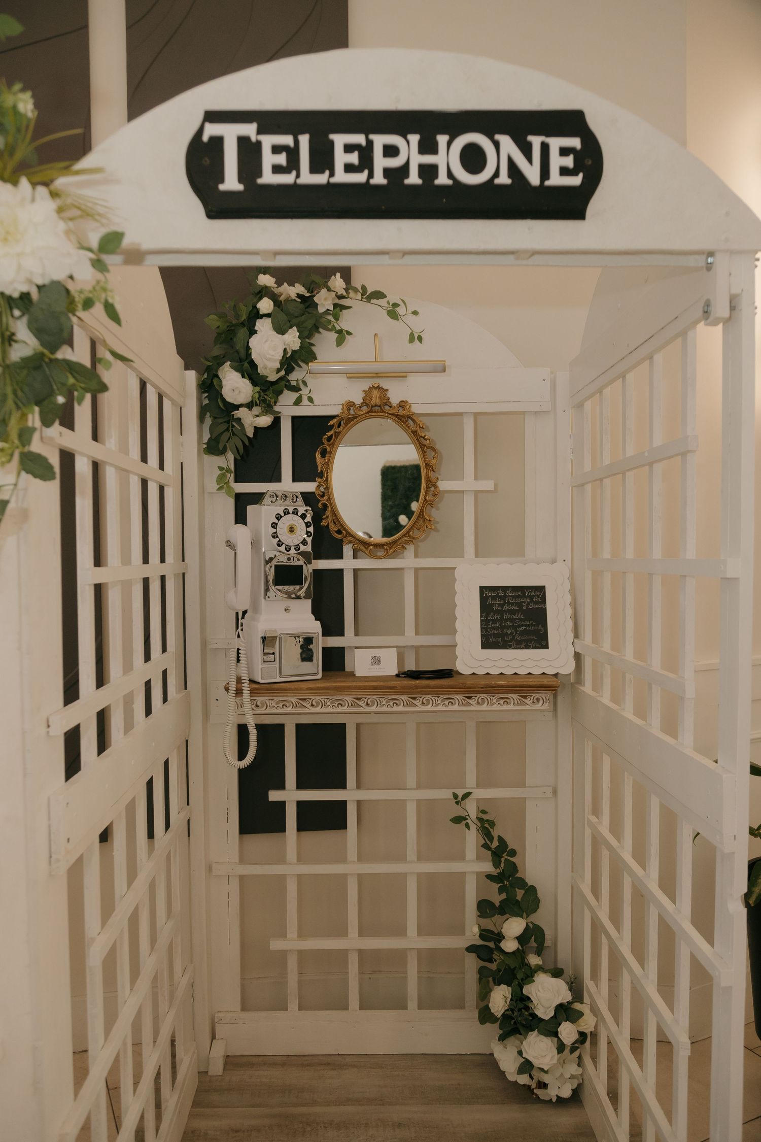 White vintage telephone booth decorated with white roses and greenery, featuring a gold mirror and rotary phone inside.
