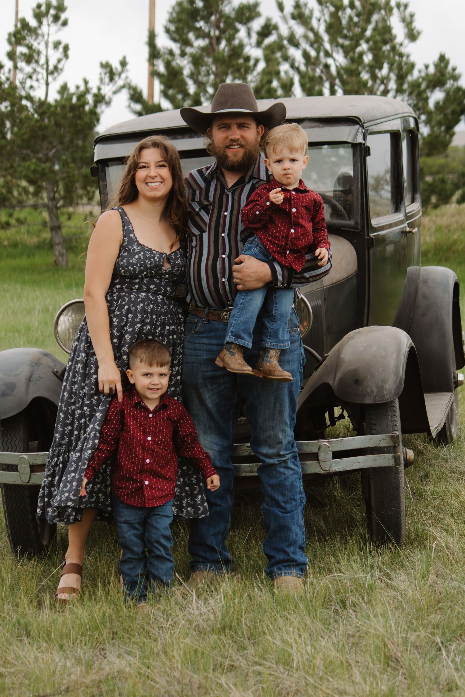 Family in coordinated outfits poses by vintage car in rustic outdoor setting.