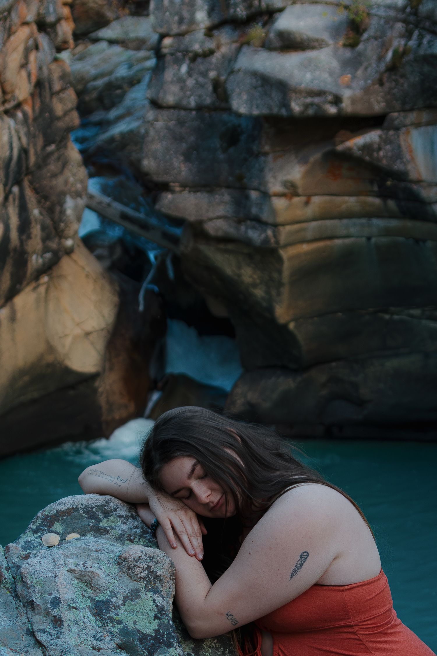 Person in red swimsuit rests on rocky edge beside turquoise mountain pool with dramatic cliff walls.