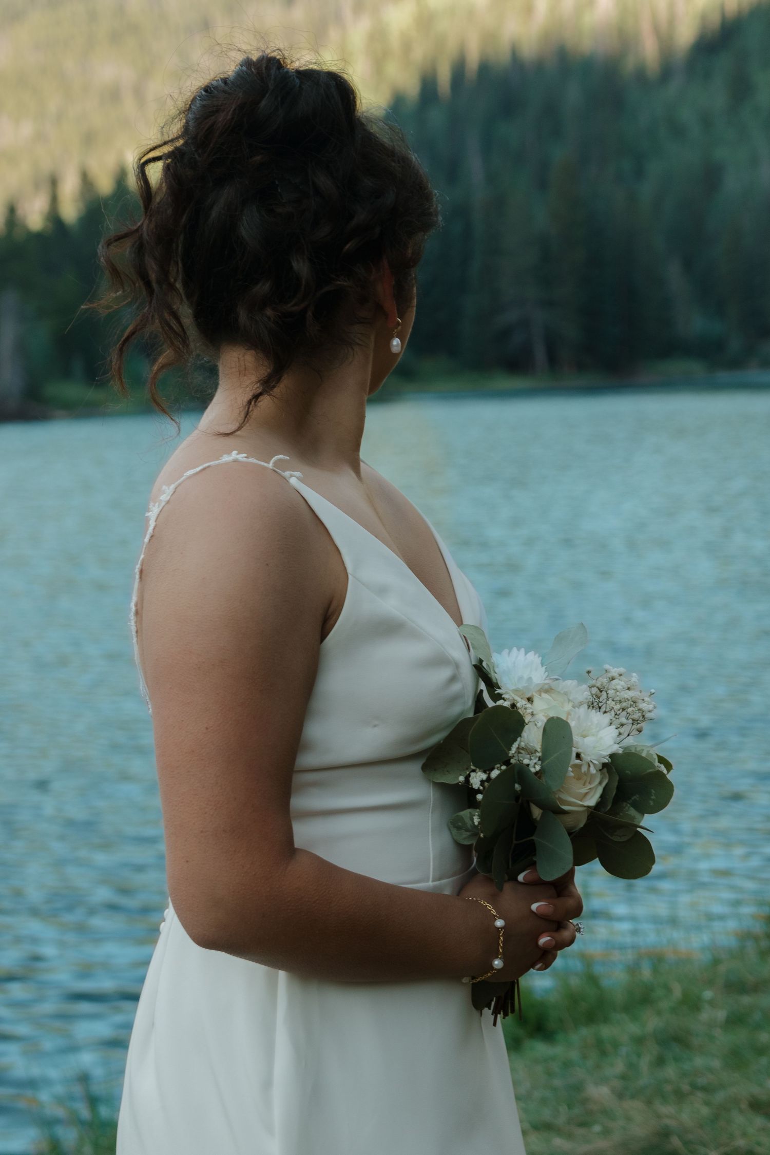 A person in a white wedding dress holds a small bouquet against a scenic mountain lake backdrop.