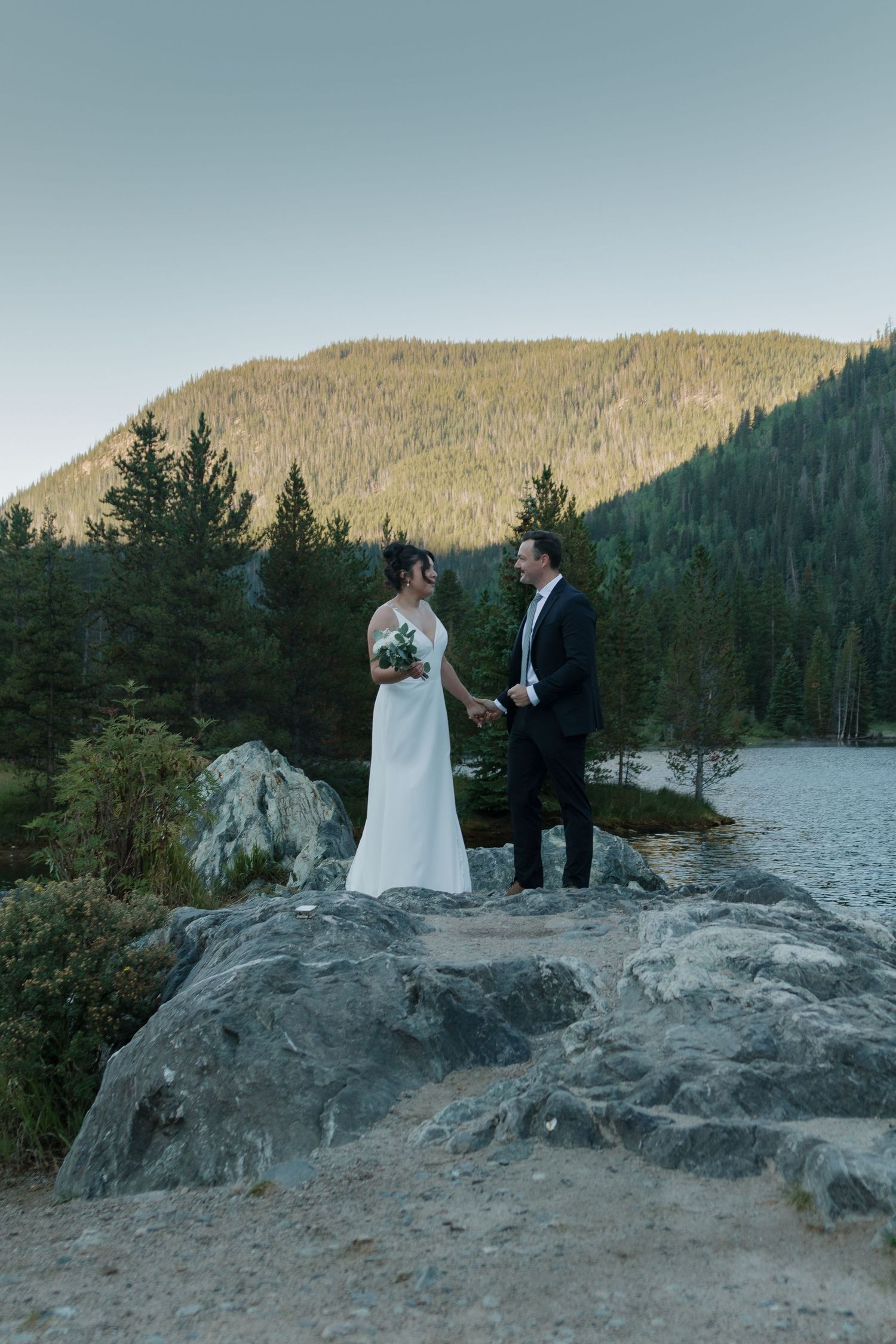 Newlyweds sharing intimate moment on rocky outcrop overlooking mountain lake at sunset.
