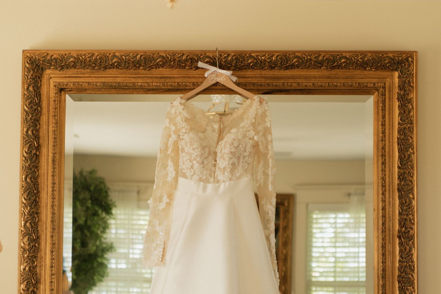 A bridal gown with delicate lace details reflects beautifully in a gold-framed mirror.