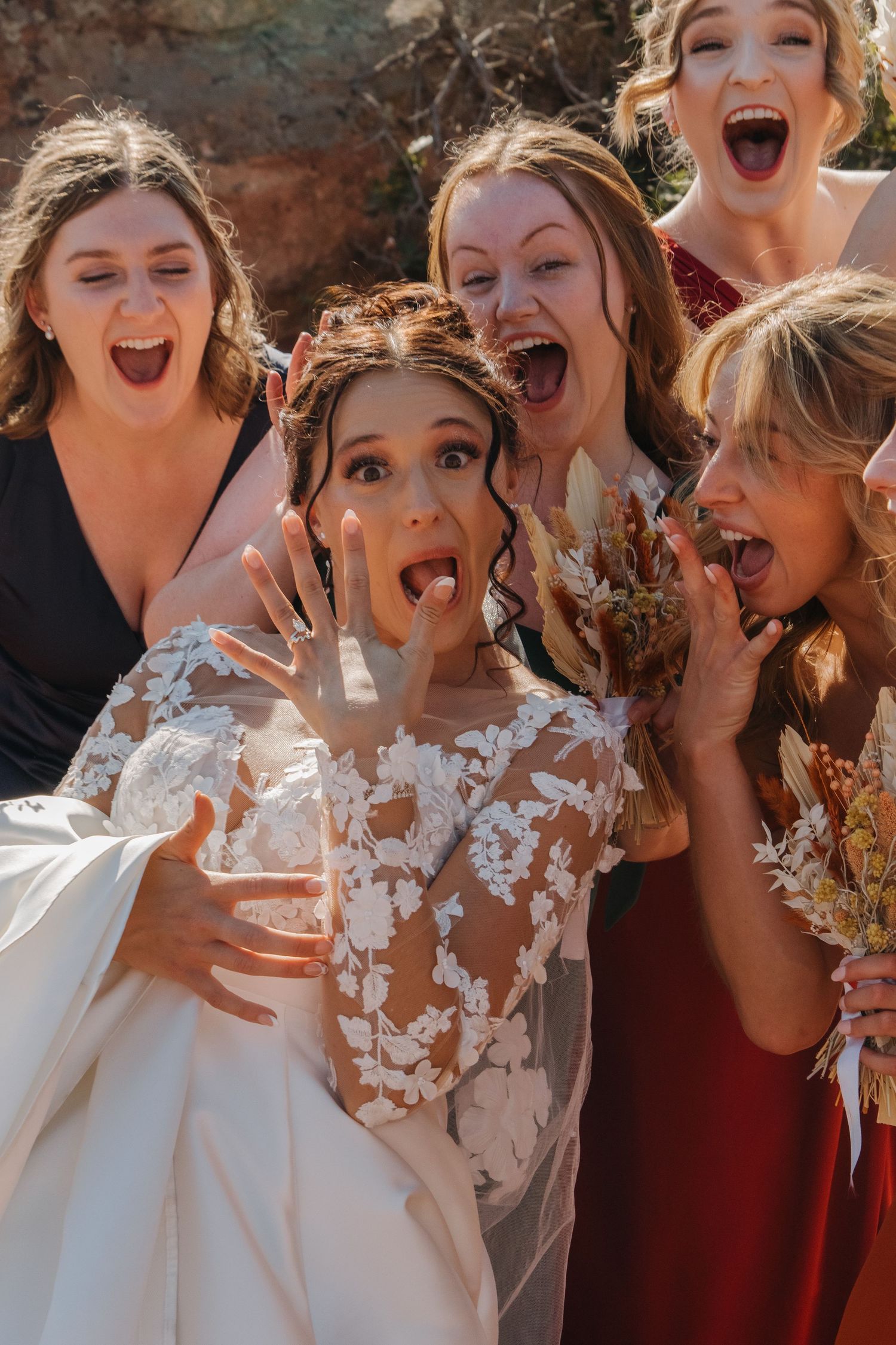 Wedding party members share joyful reactions against a rustic brick wall backdrop.