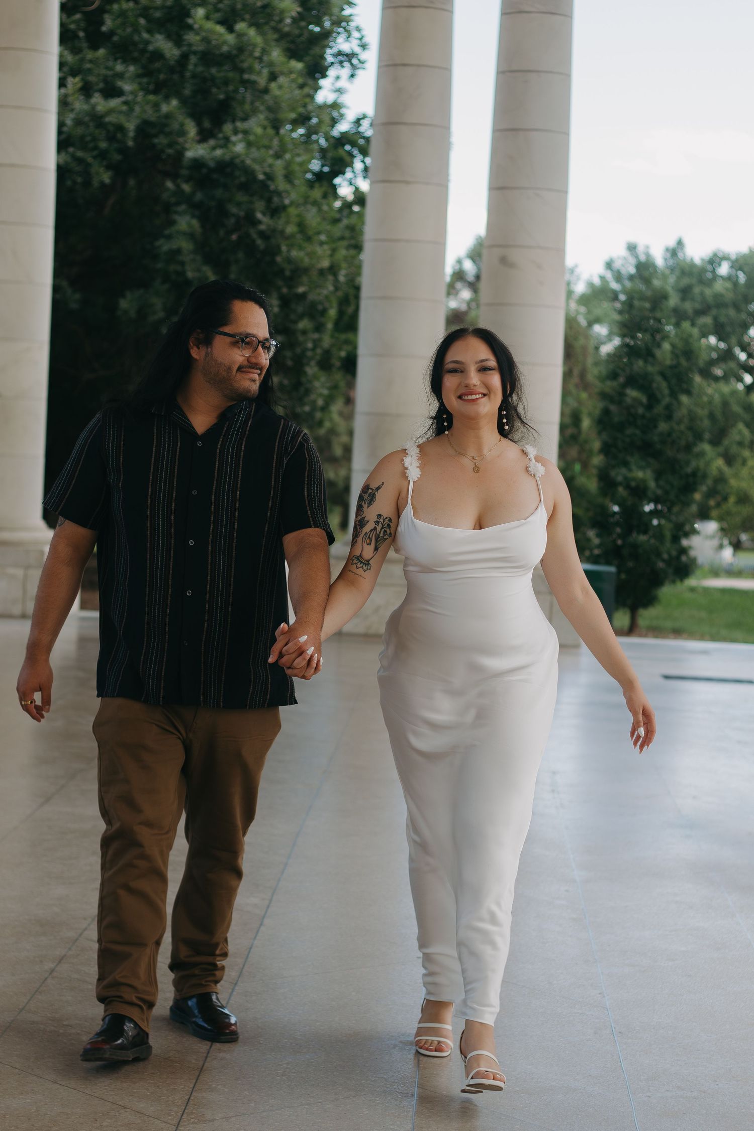 A couple walks together under white columns, with one wearing a black striped shirt and the other in a white dress.
