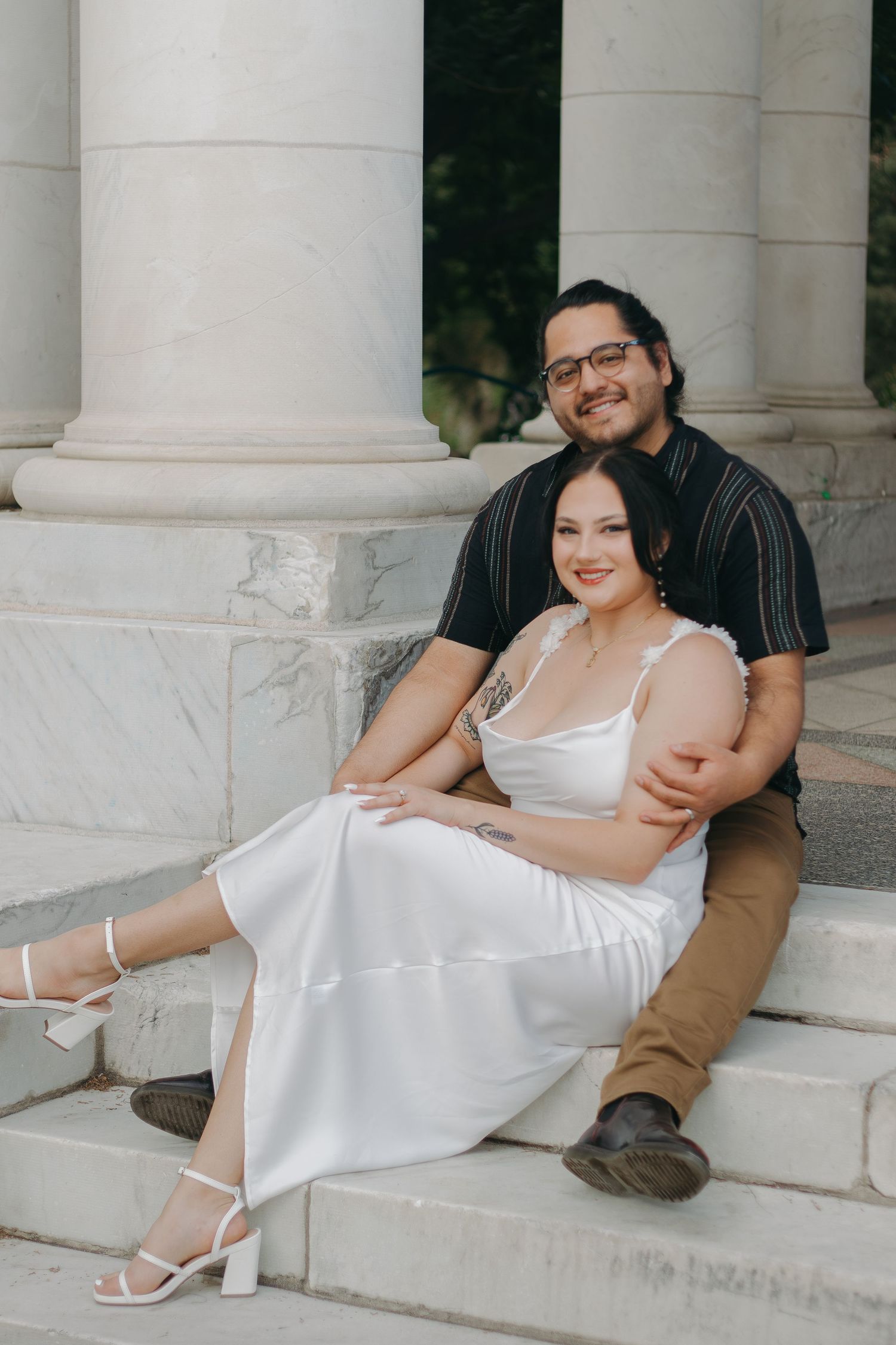 Couple sitting together on white stone steps in formal attire.