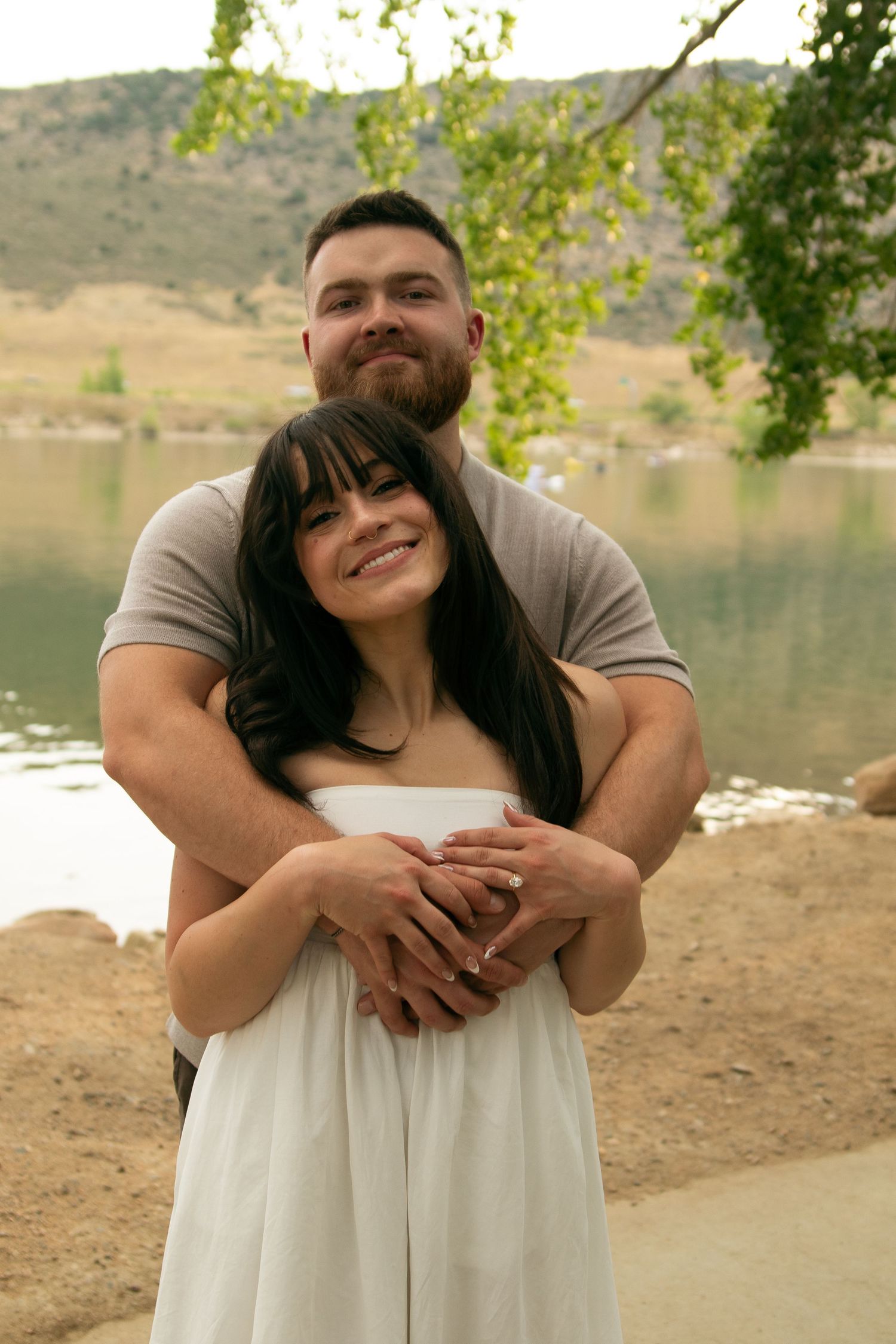 A couple embraces while posing for engagement photos by a scenic lakeside.