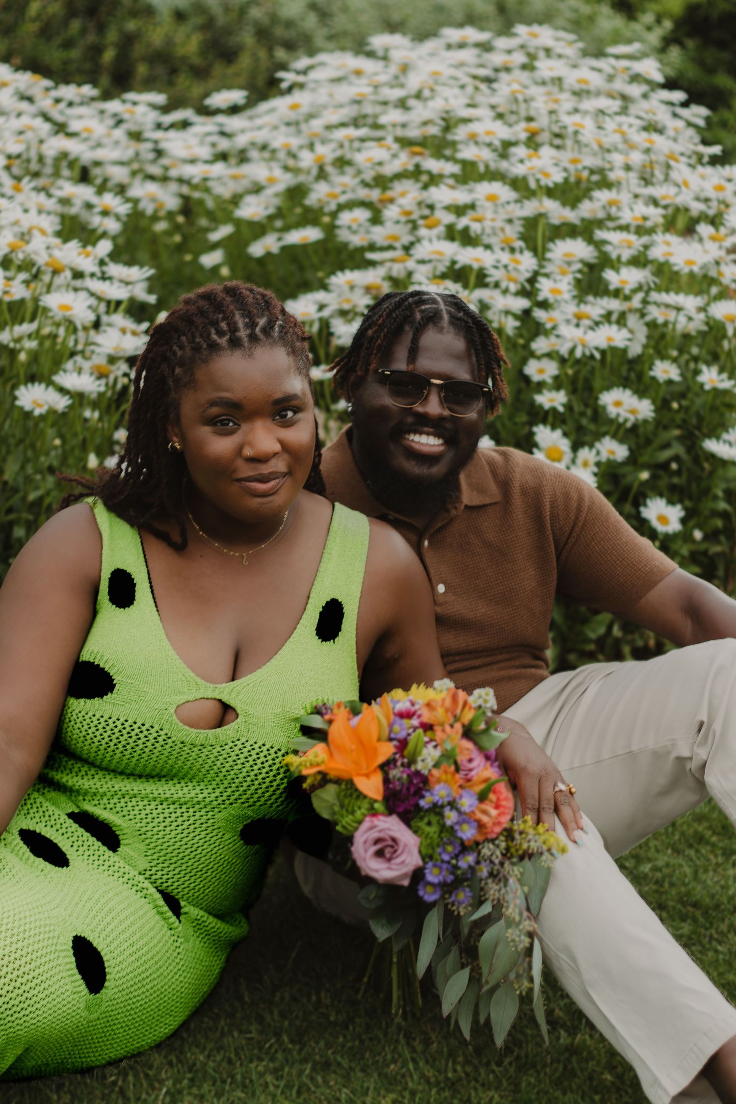 A couple poses together in a daisy field with a colorful bouquet of flowers.
