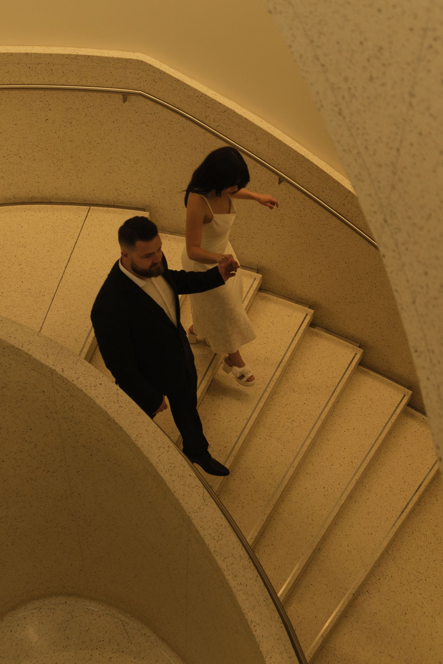 Bird's eye view sequence of figures descending curved white marble stairs.