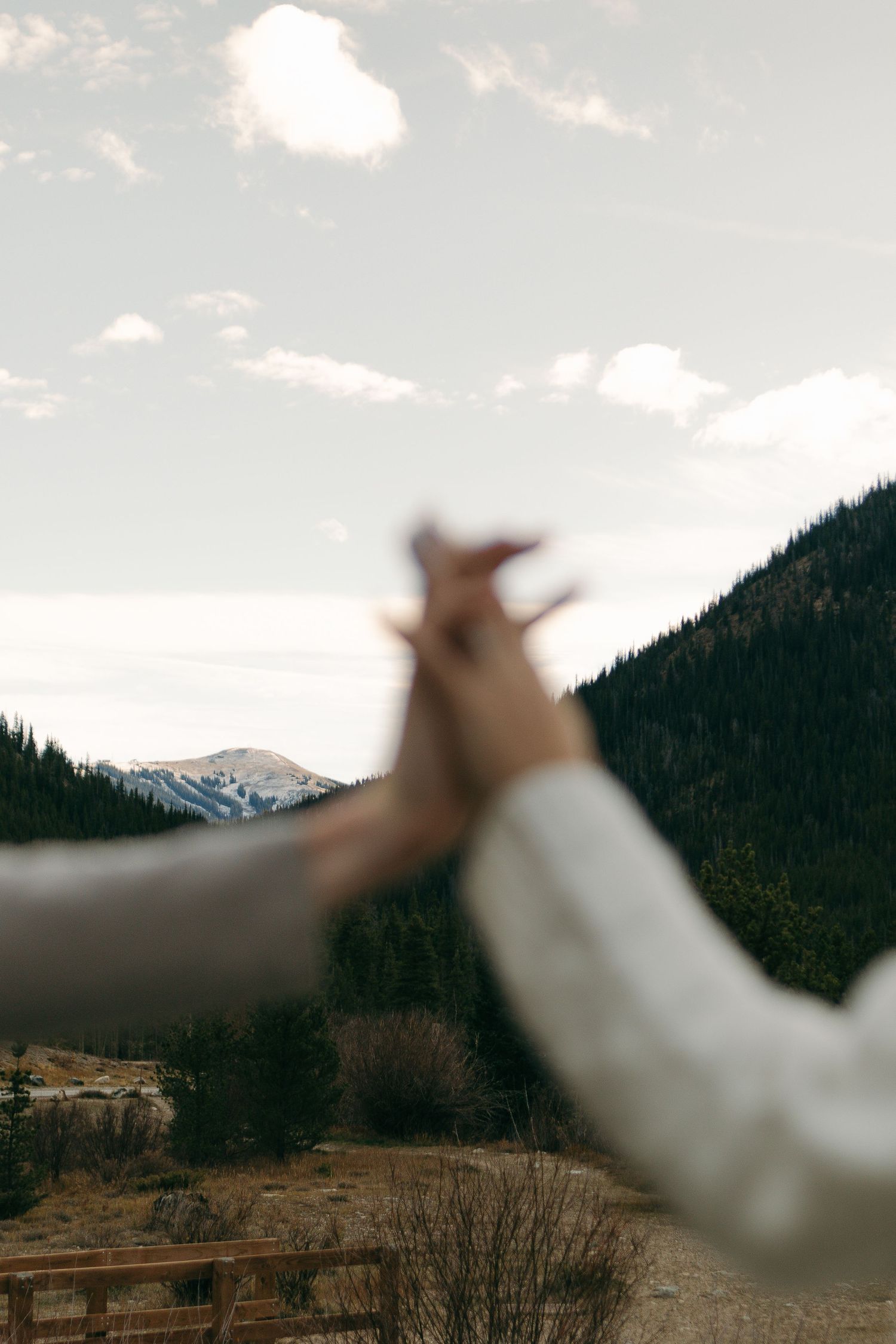 Blurred hand gestures frame a scenic mountain landscape with evergreen forests and snow-capped peaks in the distance.