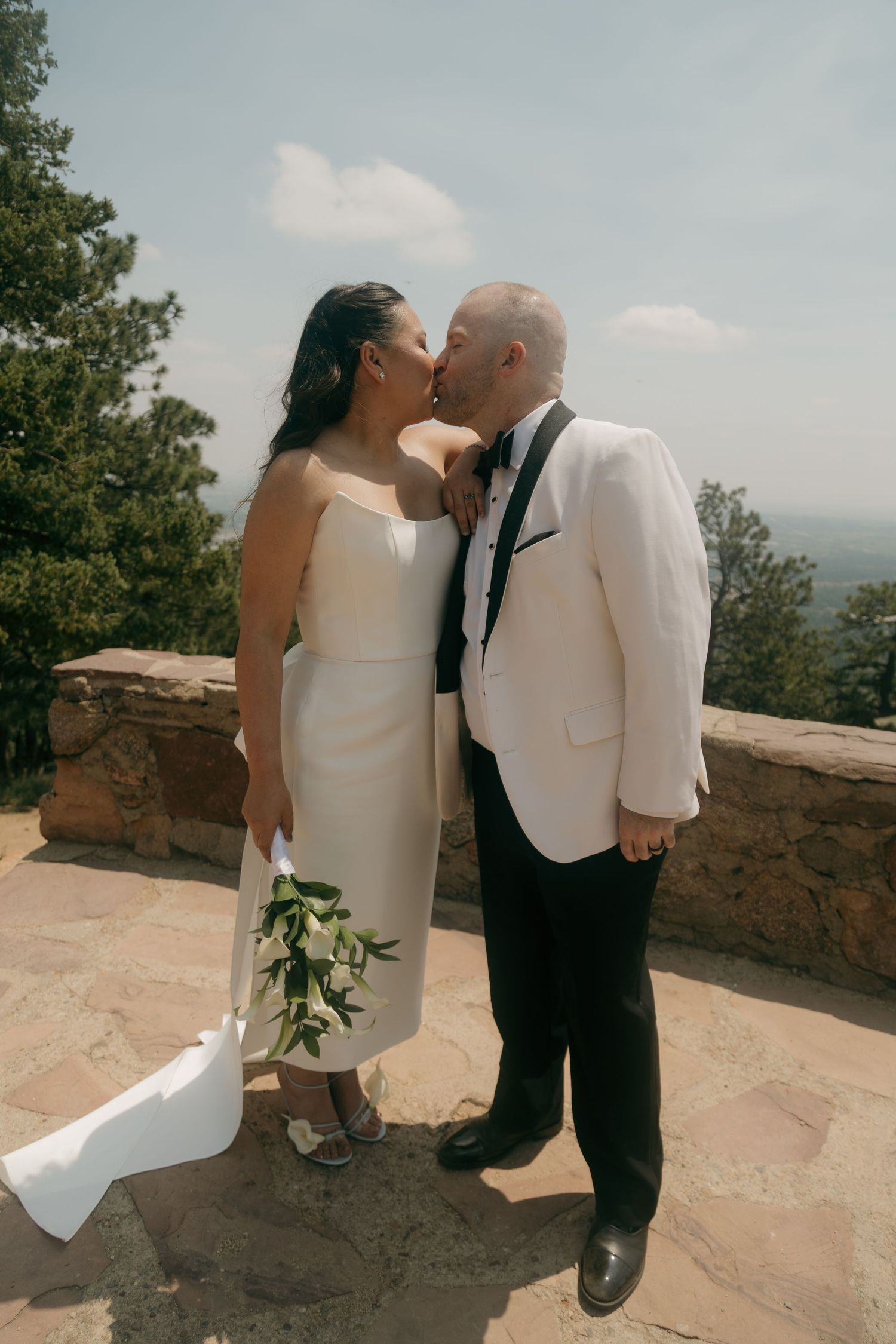Newlyweds share a romantic kiss on a stone terrace overlooking a scenic landscape, bride holding a white floral bouquet.