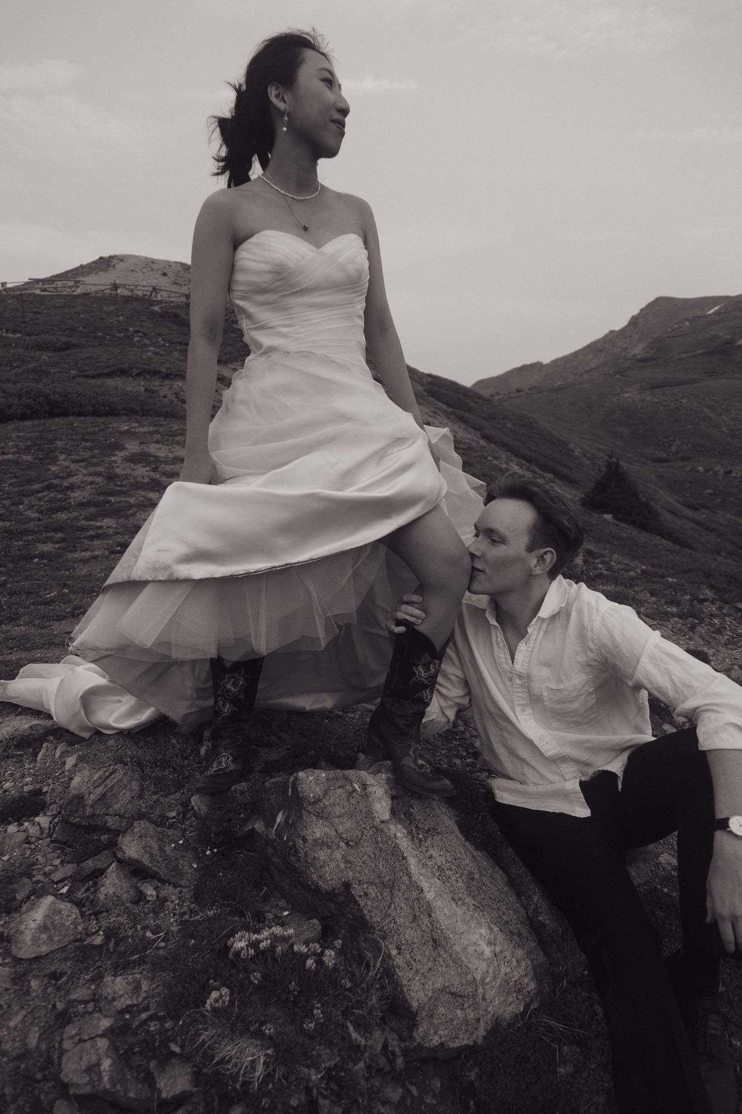 Black and white photo of bride seated on rock with groom gazing up at her in mountain setting.