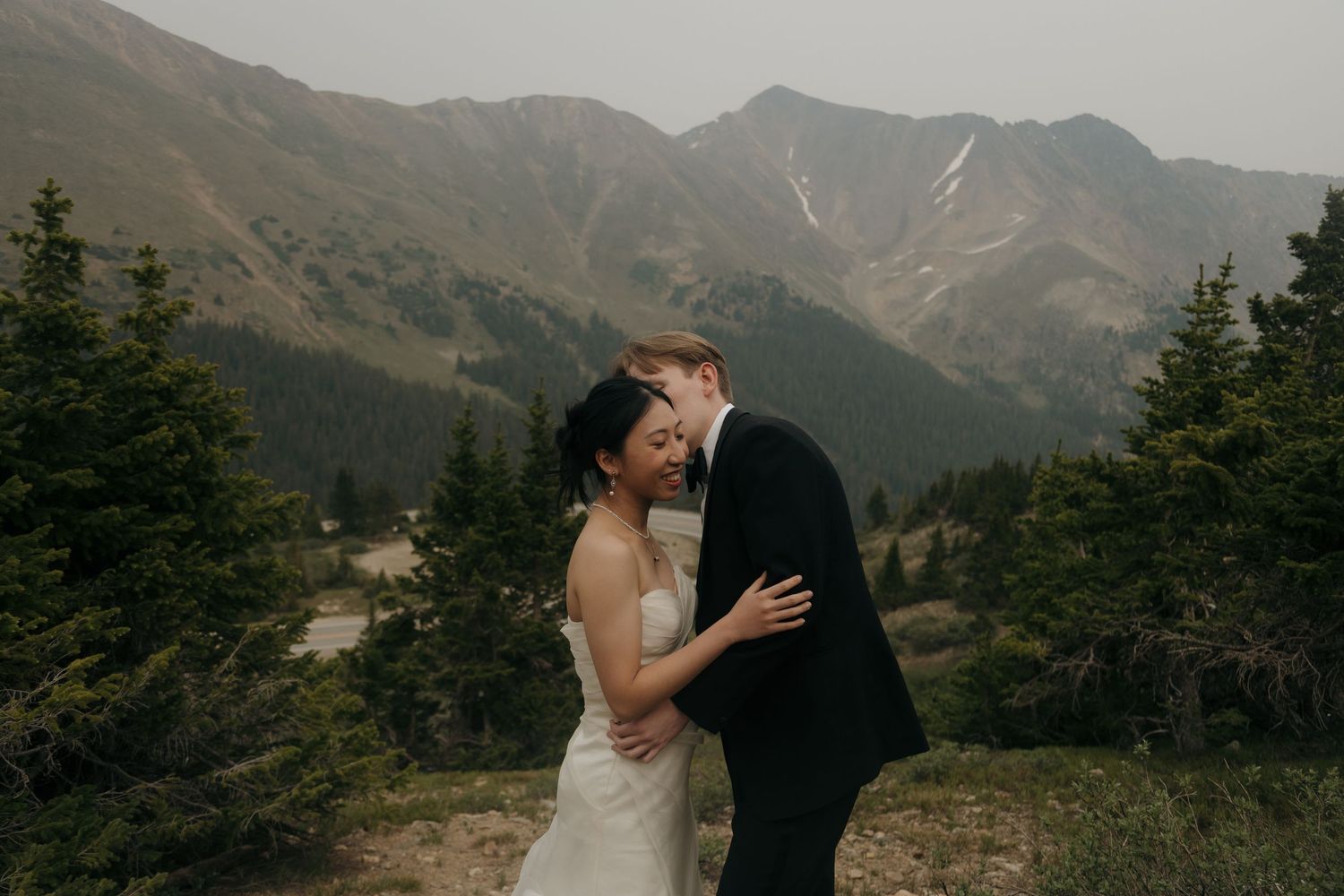 Couple in wedding attire embracing with misty mountain peaks and evergreen trees in the background.
