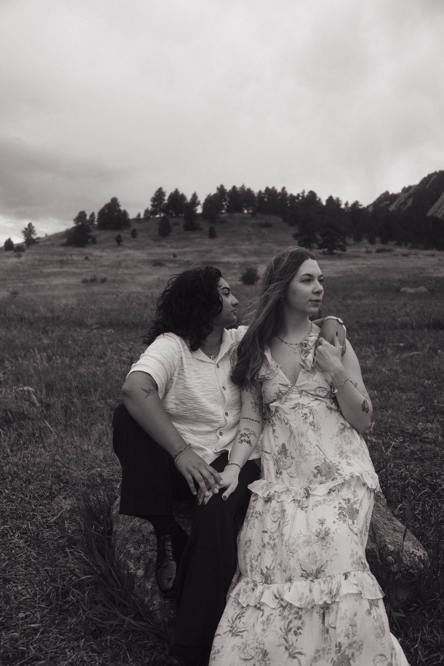 Black and white photo of a couple sitting together in a grassy field with trees and hills in the background.