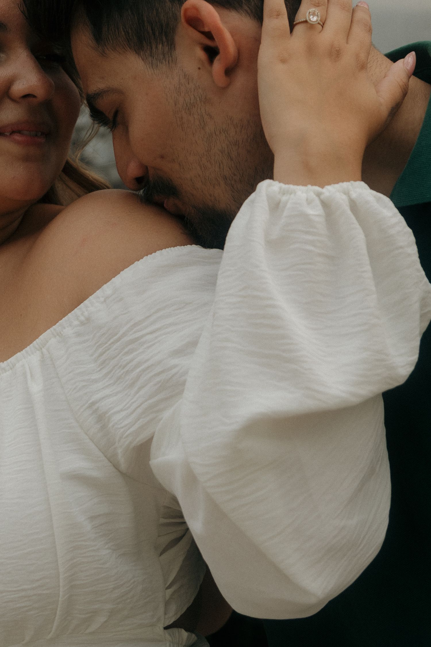 Couple embracing tenderly, woman in white off-shoulder dress with engagement ring visible on her hand resting on mans neck.
