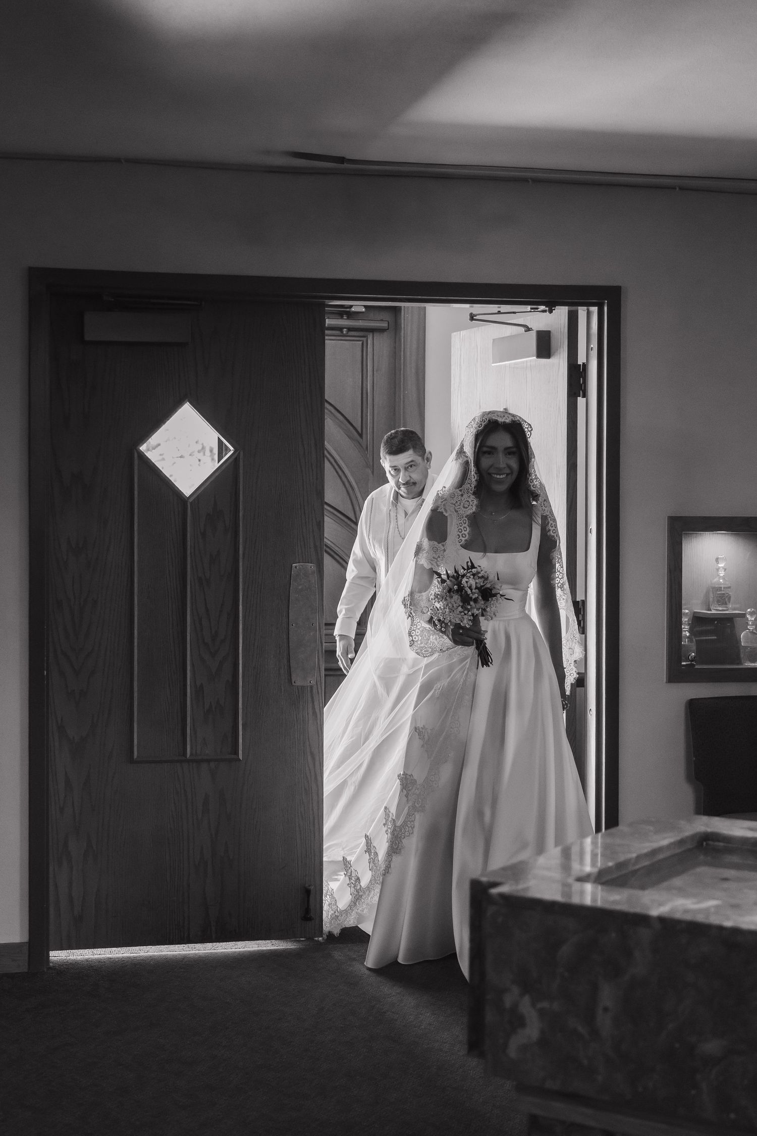 Bride and groom entering church doorway, bride smiling in white gown holding bouquet, black and white wedding photo.