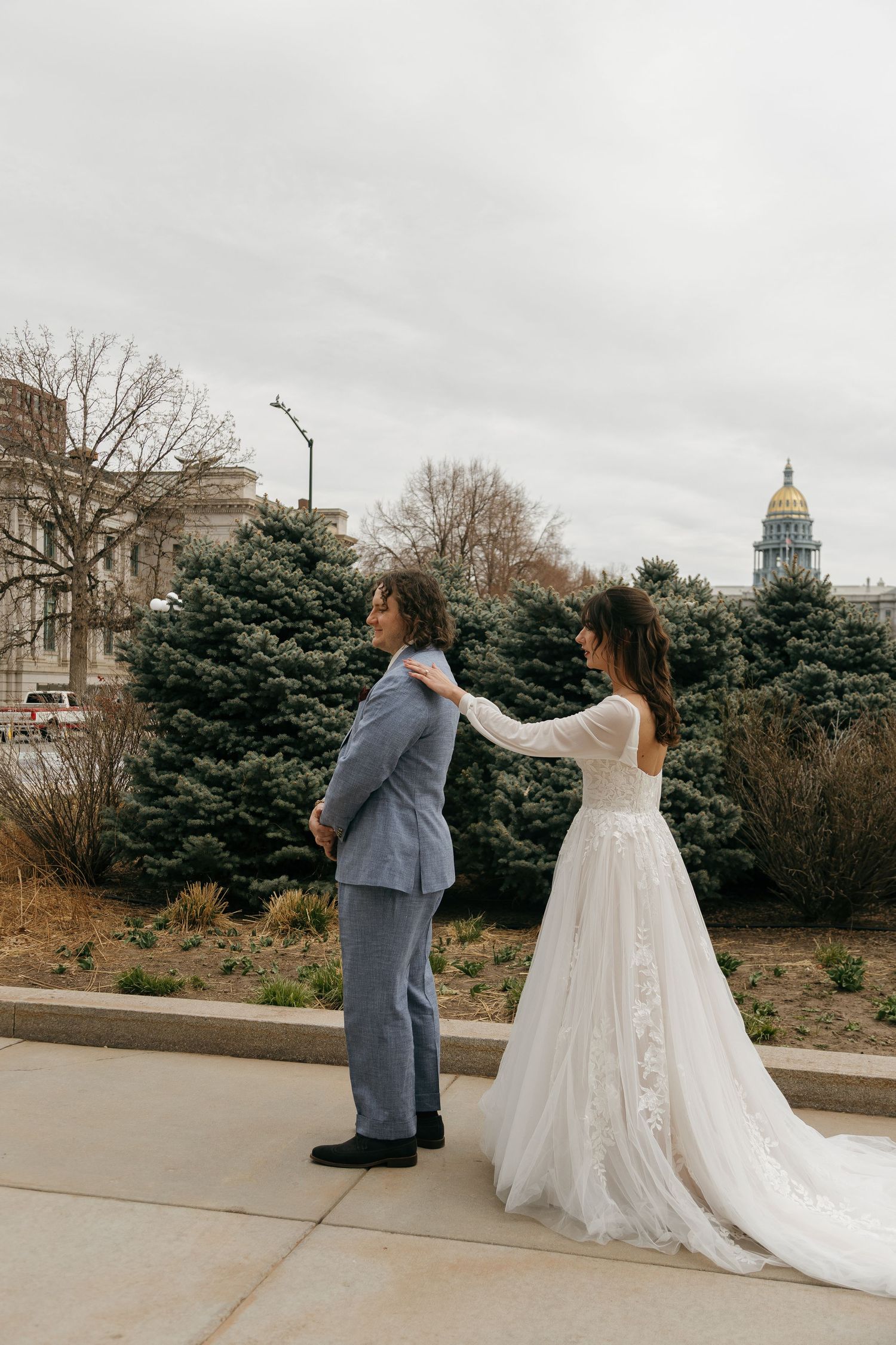A couple in wedding attire poses outside with a capitol dome visible in the background on an overcast day.