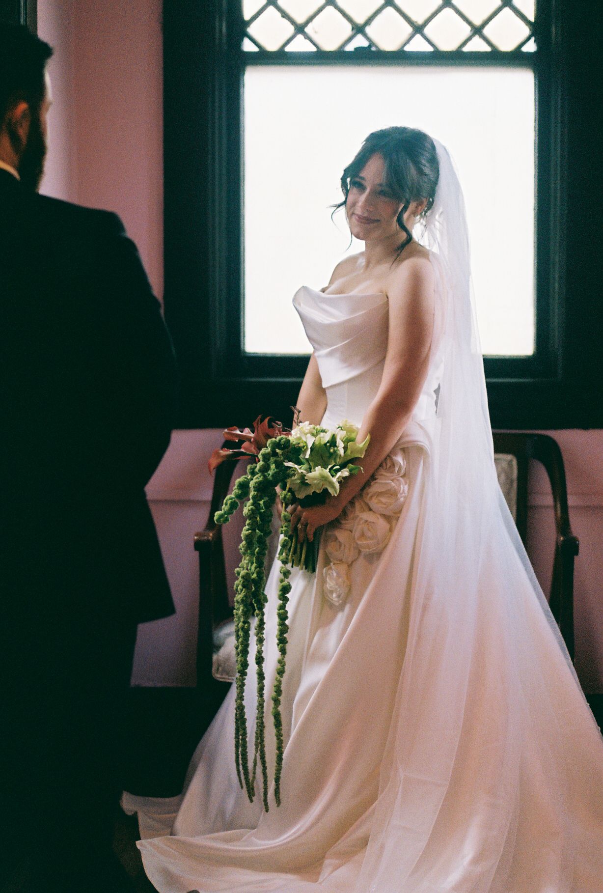 Bride in flowing white gown holds cascading bouquet near window with diamond pattern.