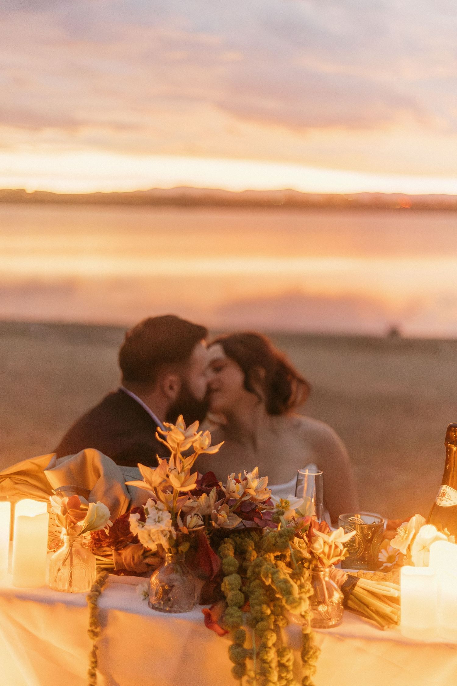 A romantic sunset dinner table with flowers and lanterns on a beach.