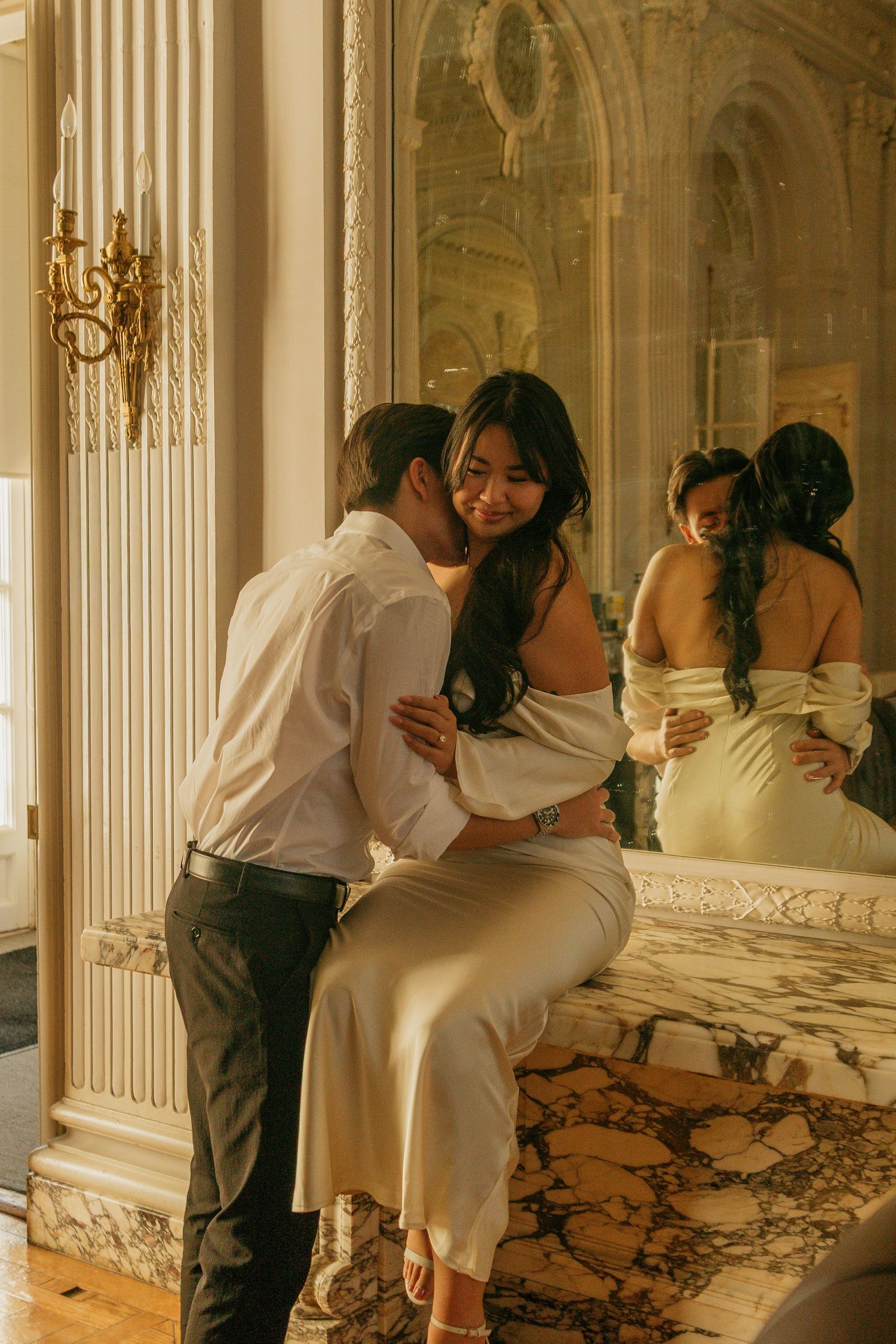 A romantic moment between a couple in formal attire reflected in an ornate mirror with marble and gold decor.
