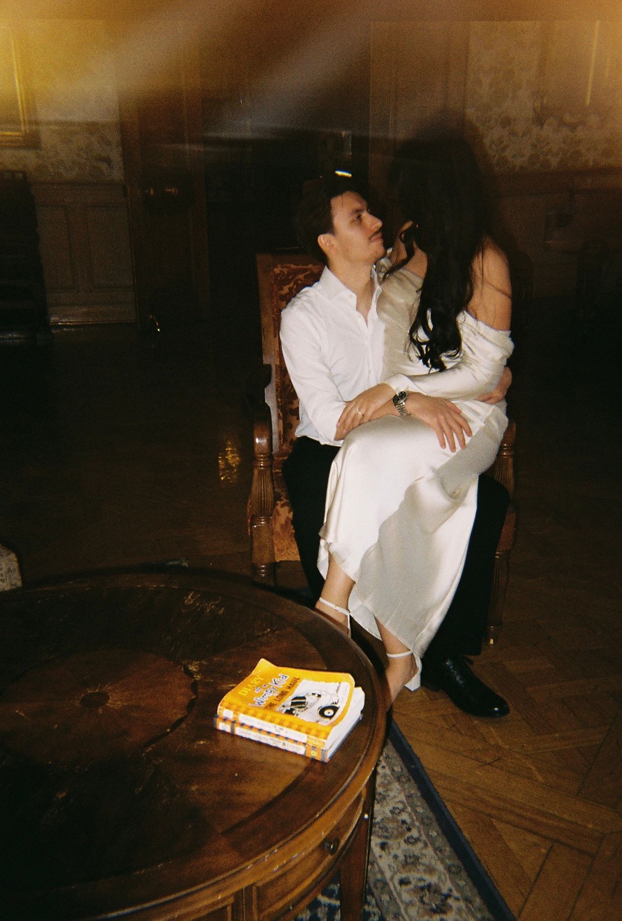 A romantic couple in white clothing shares an intimate moment in a dimly lit vintage room with a book on table.